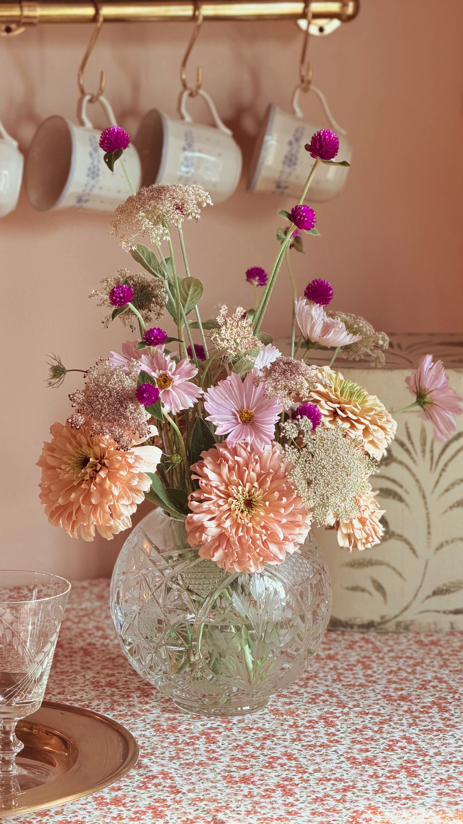 A bouquet of mixed flowers in shades of pink, peach, and purple arranged in a Vintage Etched Crystal Rose Bowl Vase on a patterned tablecloth, with hanging white mugs on a wall in the background.