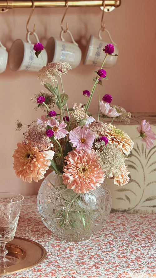 A bouquet of mixed flowers in shades of pink, peach, and purple arranged in a Vintage Etched Crystal Rose Bowl Vase on a patterned tablecloth, with hanging white mugs on a wall in the background.