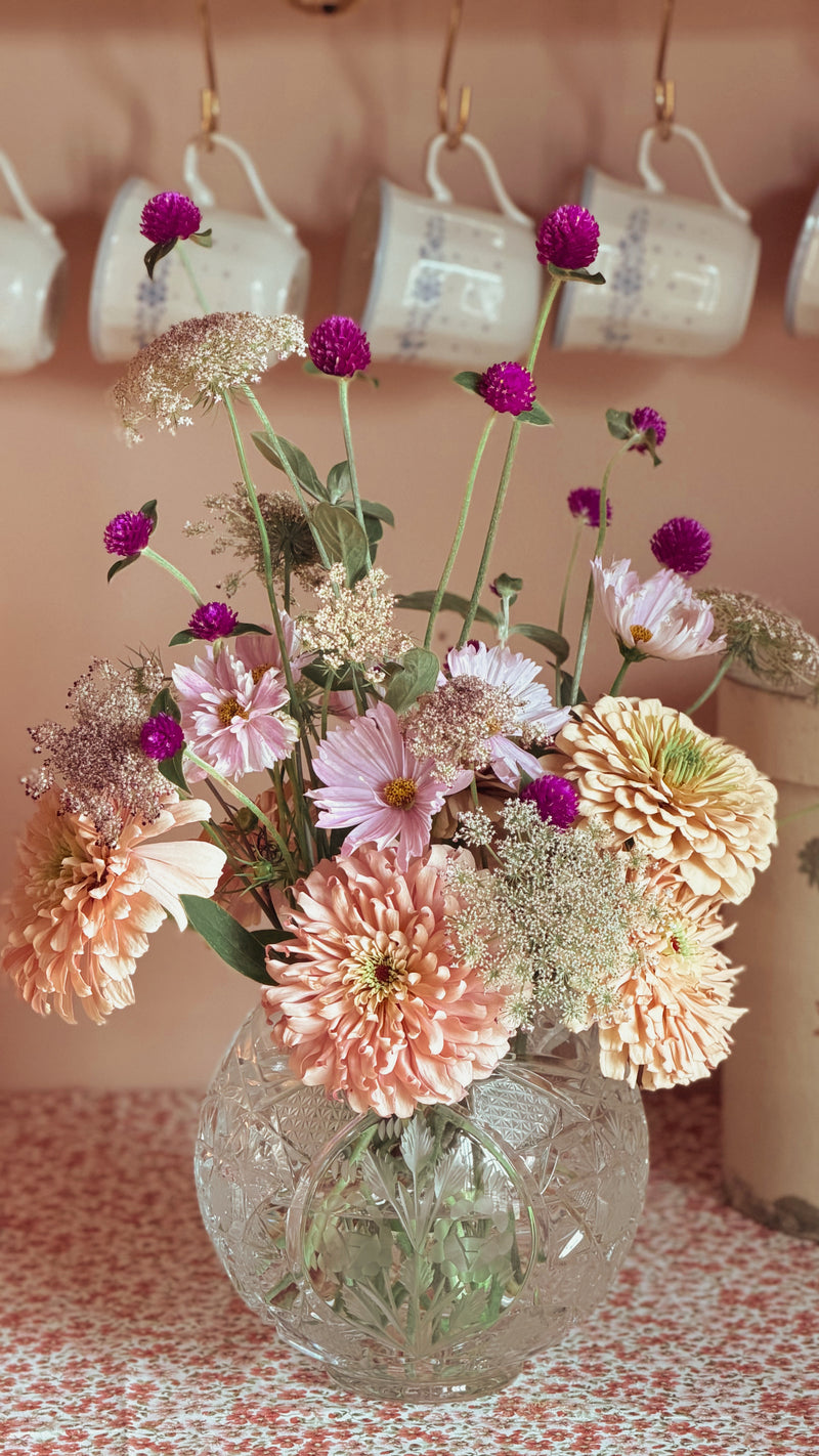 A colorful bouquet in a Vintage Etched Crystal Rose Bowl Vase, set on a patterned tablecloth with vintage teacups hanging on a pink wall in the background.