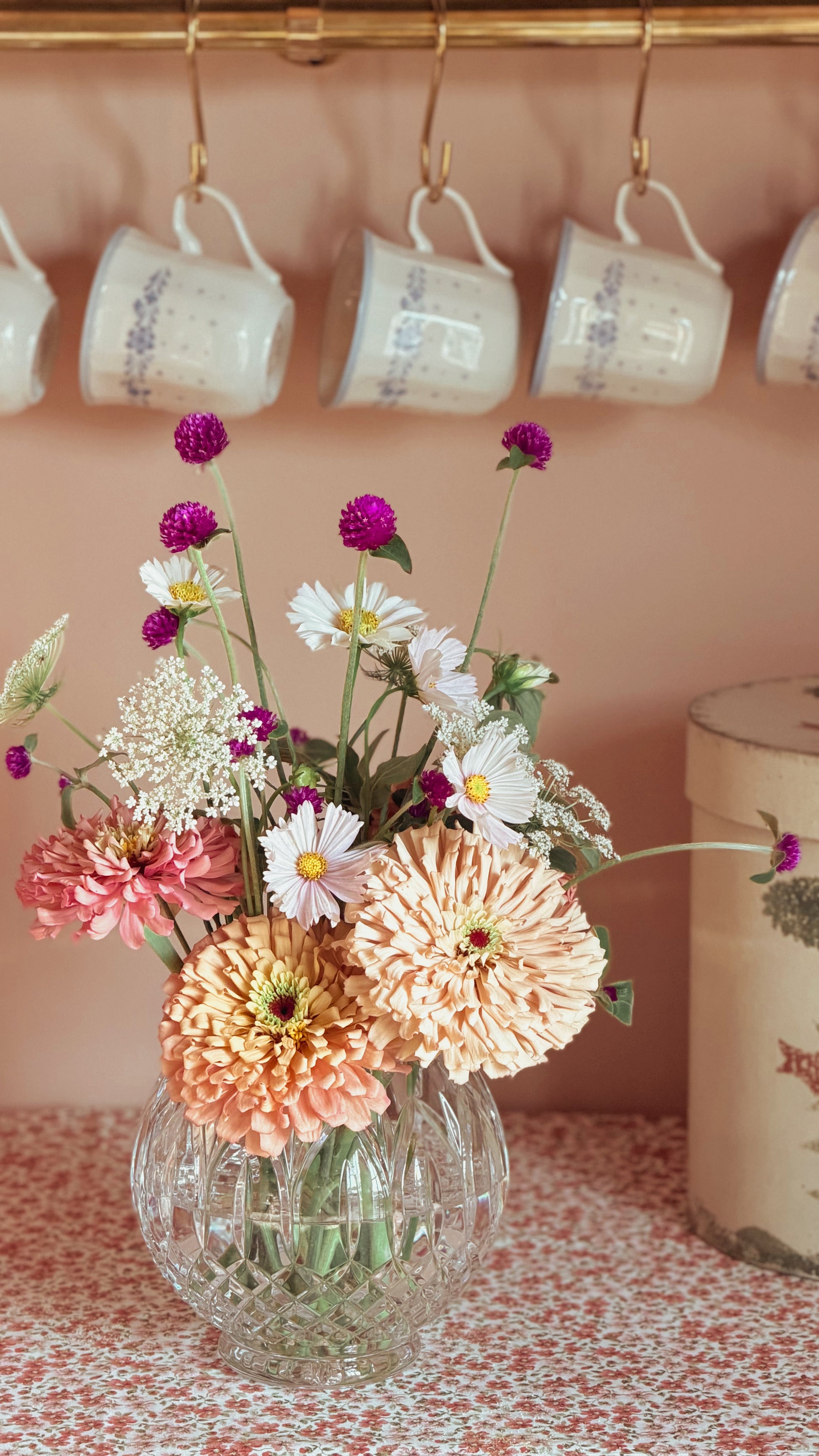 A colorful flower bouquet in a Vintage Crystal Rose Bowl Vase, set on a patterned tablecloth with a pink wall and hanging white mugs in the background.