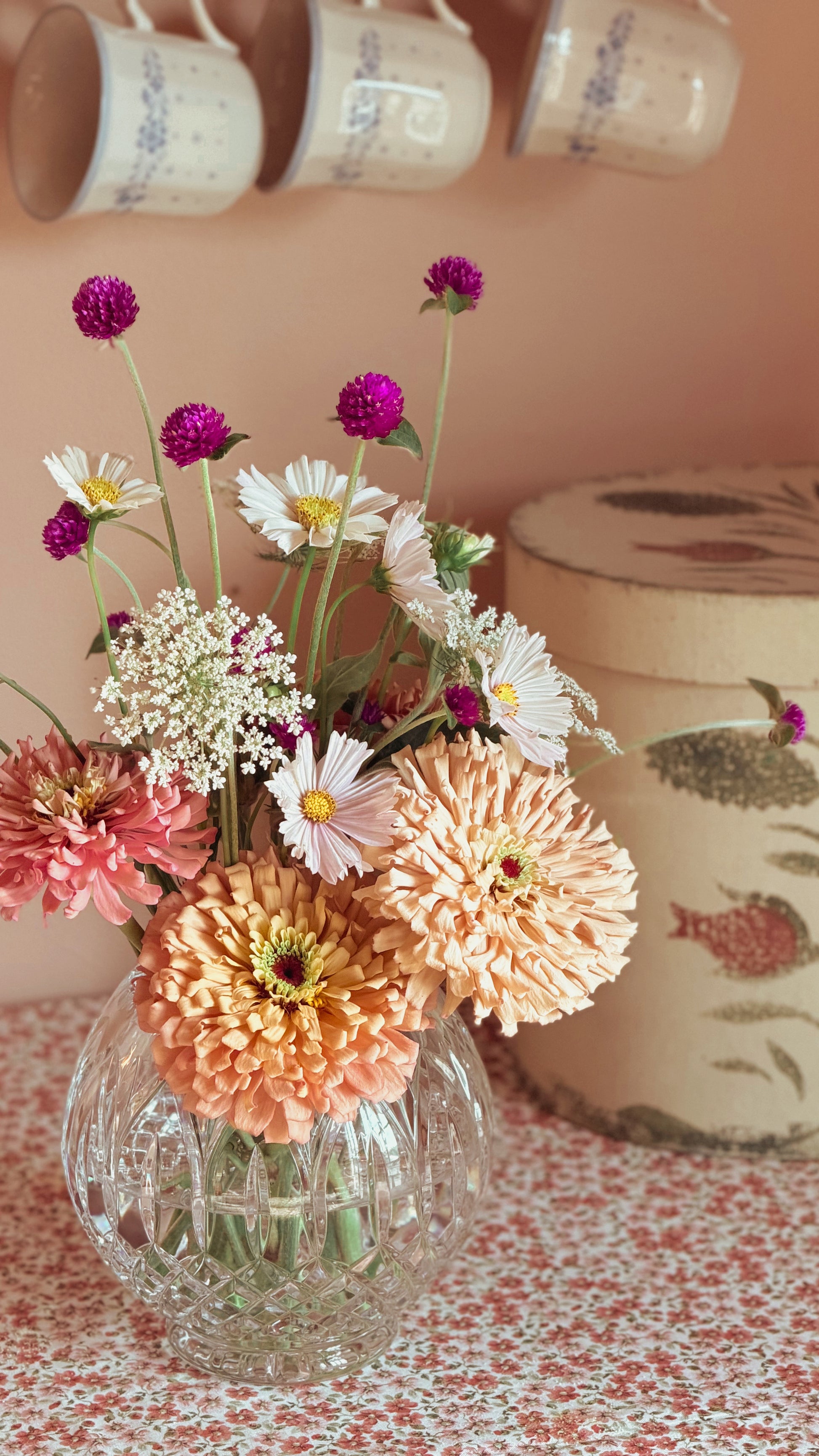 A glass vase filled with a bouquet of mixed flowers, including peach chrysanthemums, white daisies, and purple globes, set on a patterned tablecloth with a decorative tin and hanging mugs in the background.