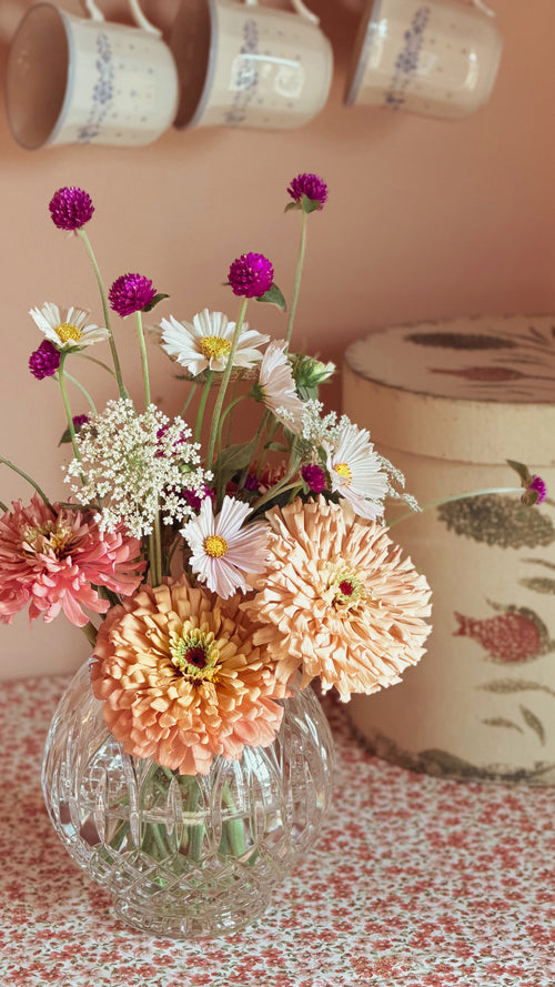A glass vase filled with a bouquet of mixed flowers, including peach chrysanthemums, white daisies, and purple globes, set on a patterned tablecloth with a decorative tin and hanging mugs in the background.