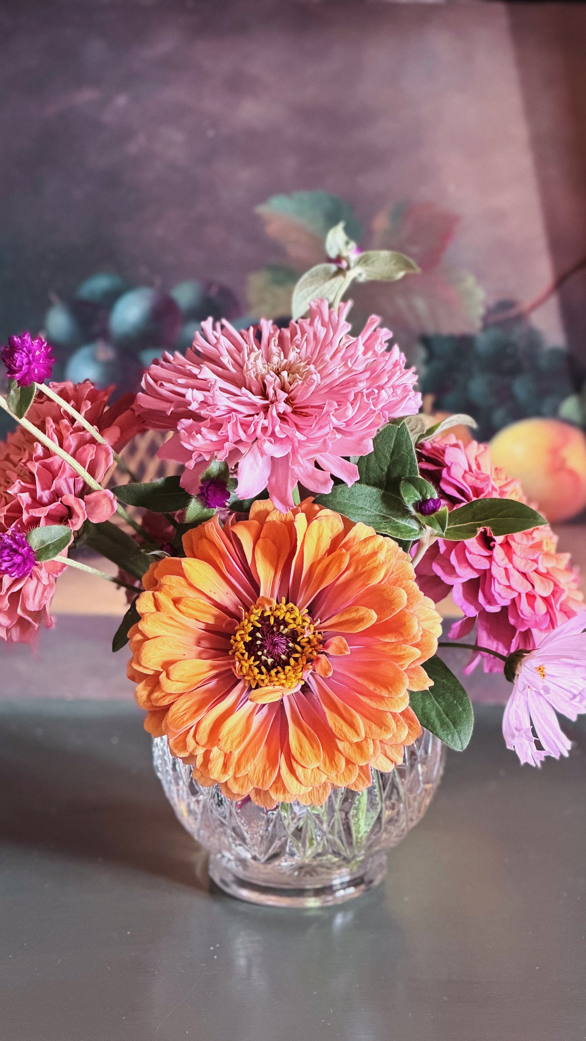 A vibrant floral arrangement featuring a prominent orange and pink zinnia flower, pink chrysanthemums, and other colorful blooms in a clear glass vase, set against a dark, painterly background.