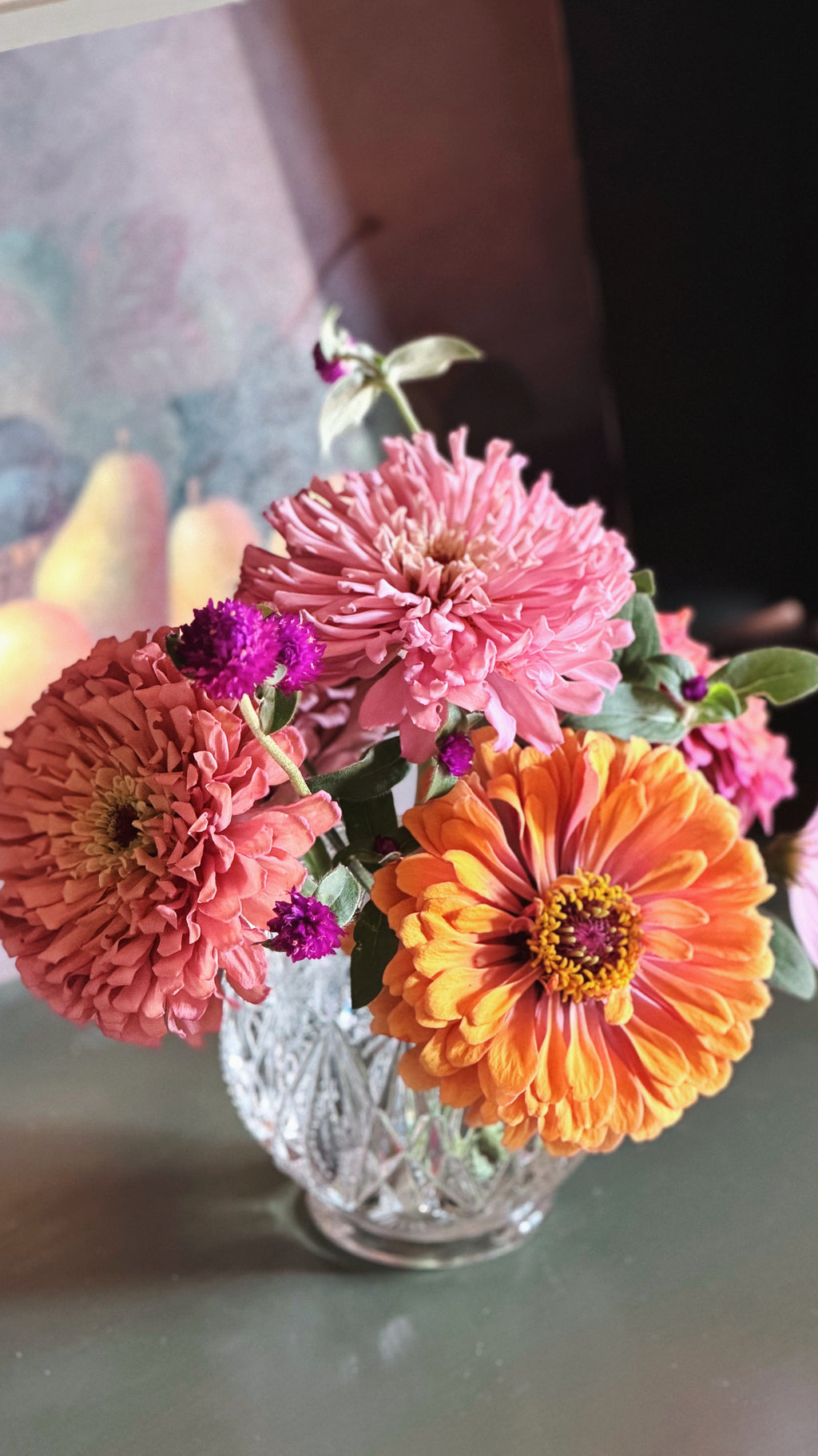 A bouquet of fresh zinnia flowers in a Vintage Avon Pressed Glass Votive Candle Holder, featuring a large pink flower, a bright orange flower, and smaller purple blossoms on a tabletop.