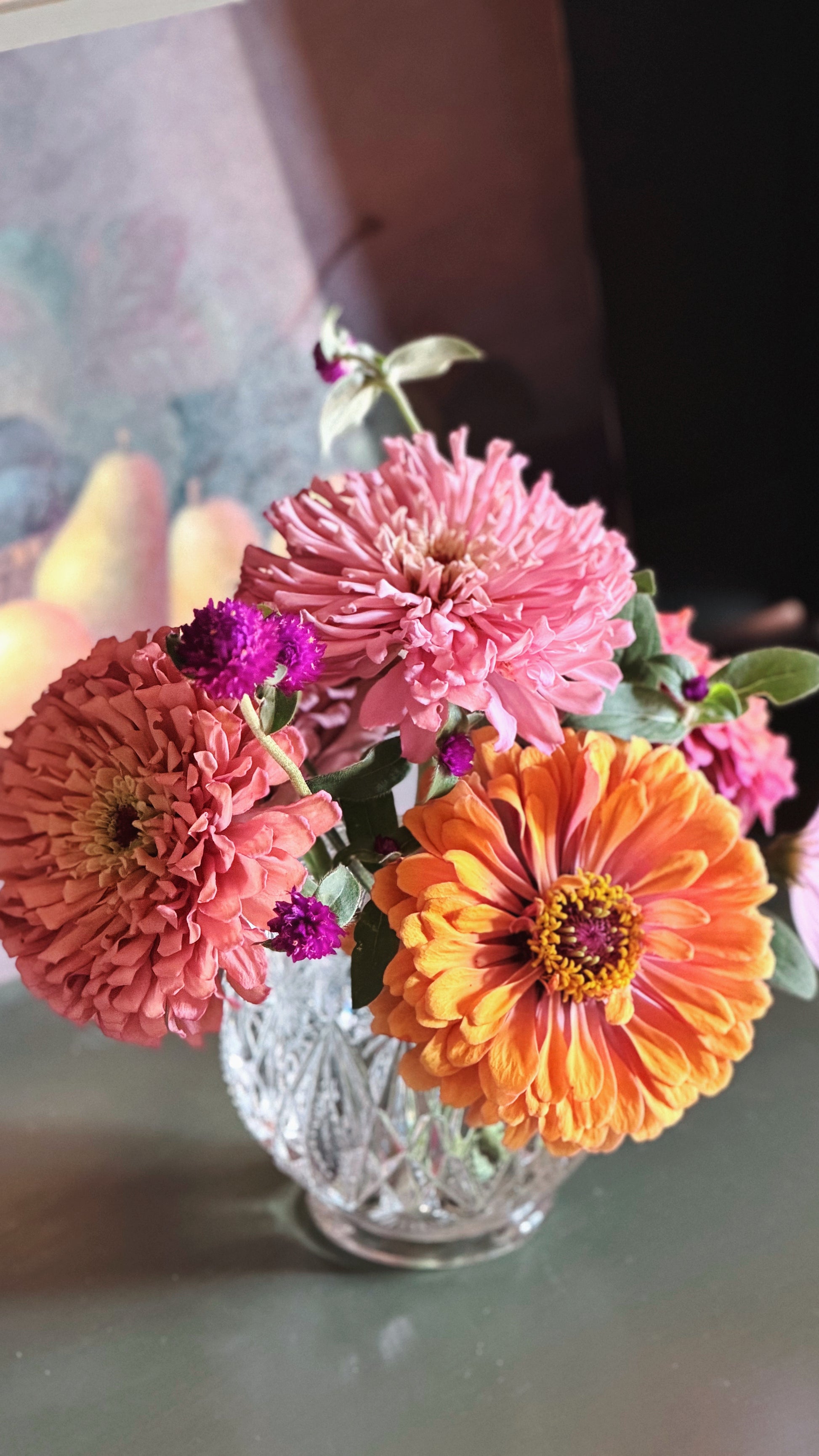 A bouquet of fresh zinnia flowers in a Vintage Avon Pressed Glass Votive Candle Holder, featuring a large pink flower, a bright orange flower, and smaller purple blossoms on a tabletop.