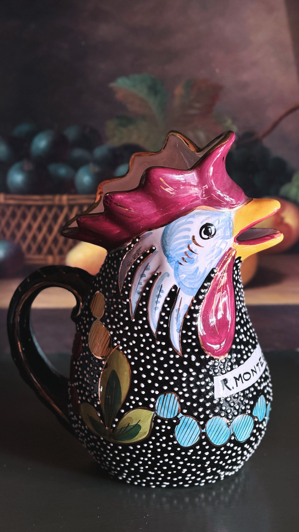 A hand-painted ceramic pitcher shaped like a rooster, featuring a black body with white polka dots, a pink and blue face, and colorful decorative accents, set against a dark, blurred background.