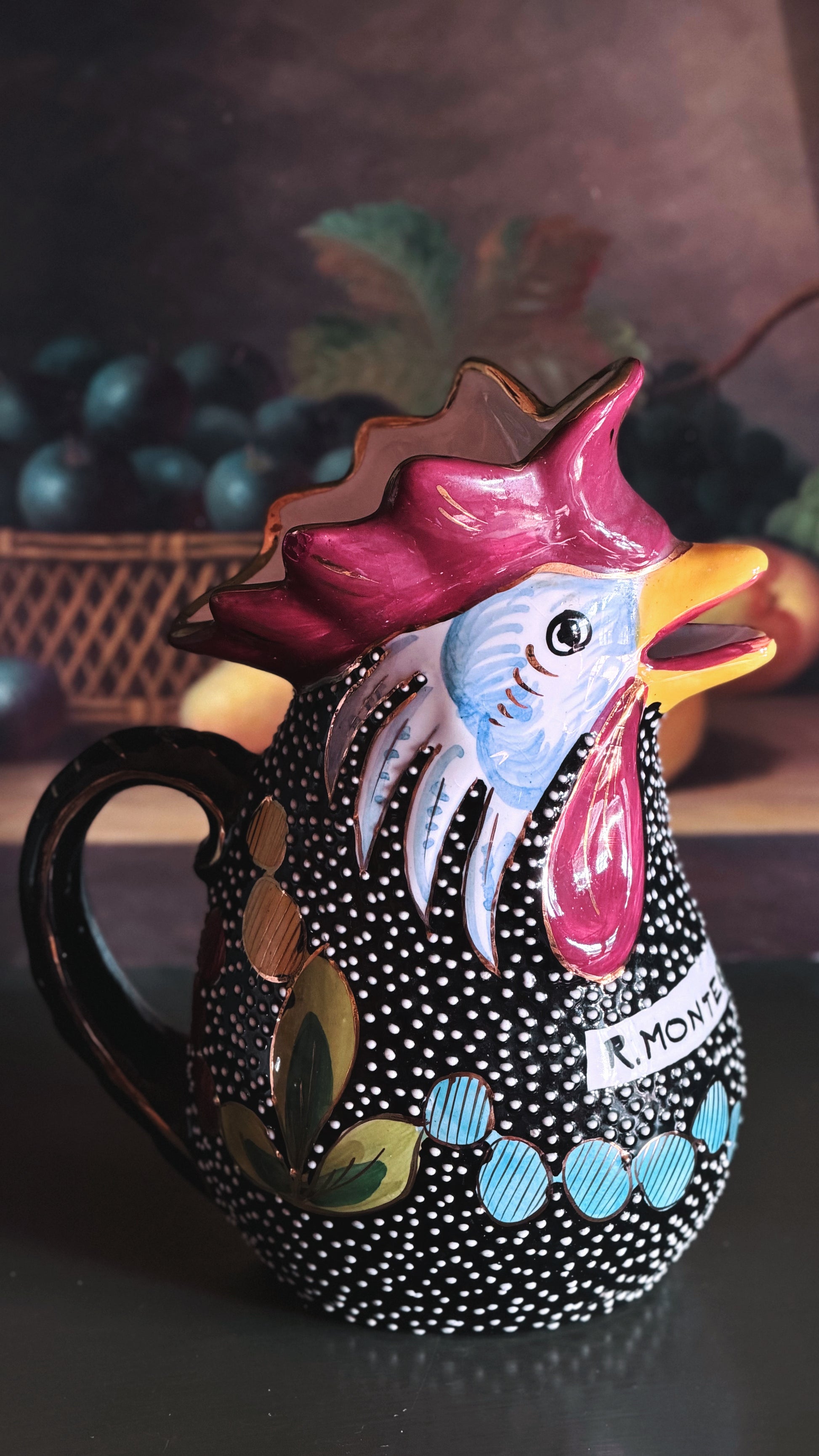 A hand-painted ceramic pitcher shaped like a rooster, featuring a black body with white polka dots, a pink and blue face, and colorful decorative accents, set against a dark, blurred background.