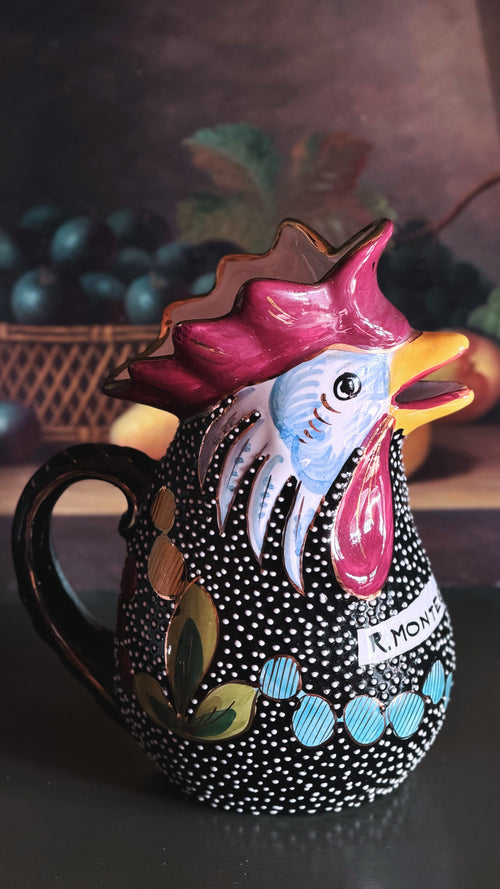 A hand-painted ceramic pitcher shaped like a rooster, featuring a black body with white polka dots, a pink and blue face, and colorful decorative accents, set against a dark, blurred background.