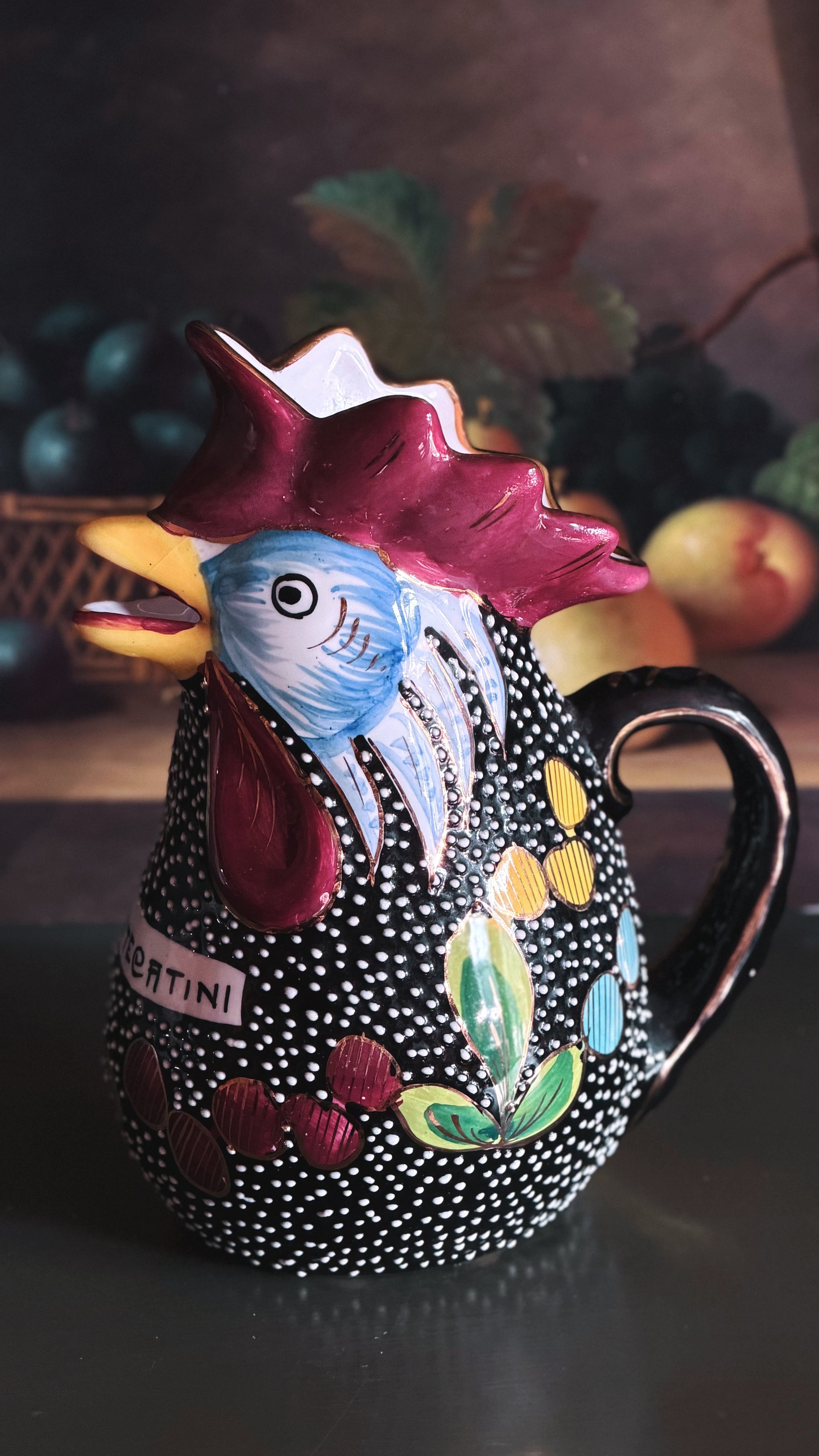 A hand-painted ceramic pitcher shaped like a rooster, featuring a black and white speckled body, a red comb, a blue face, and colorful floral accents, set against a dark rustic background with blurred fruit.