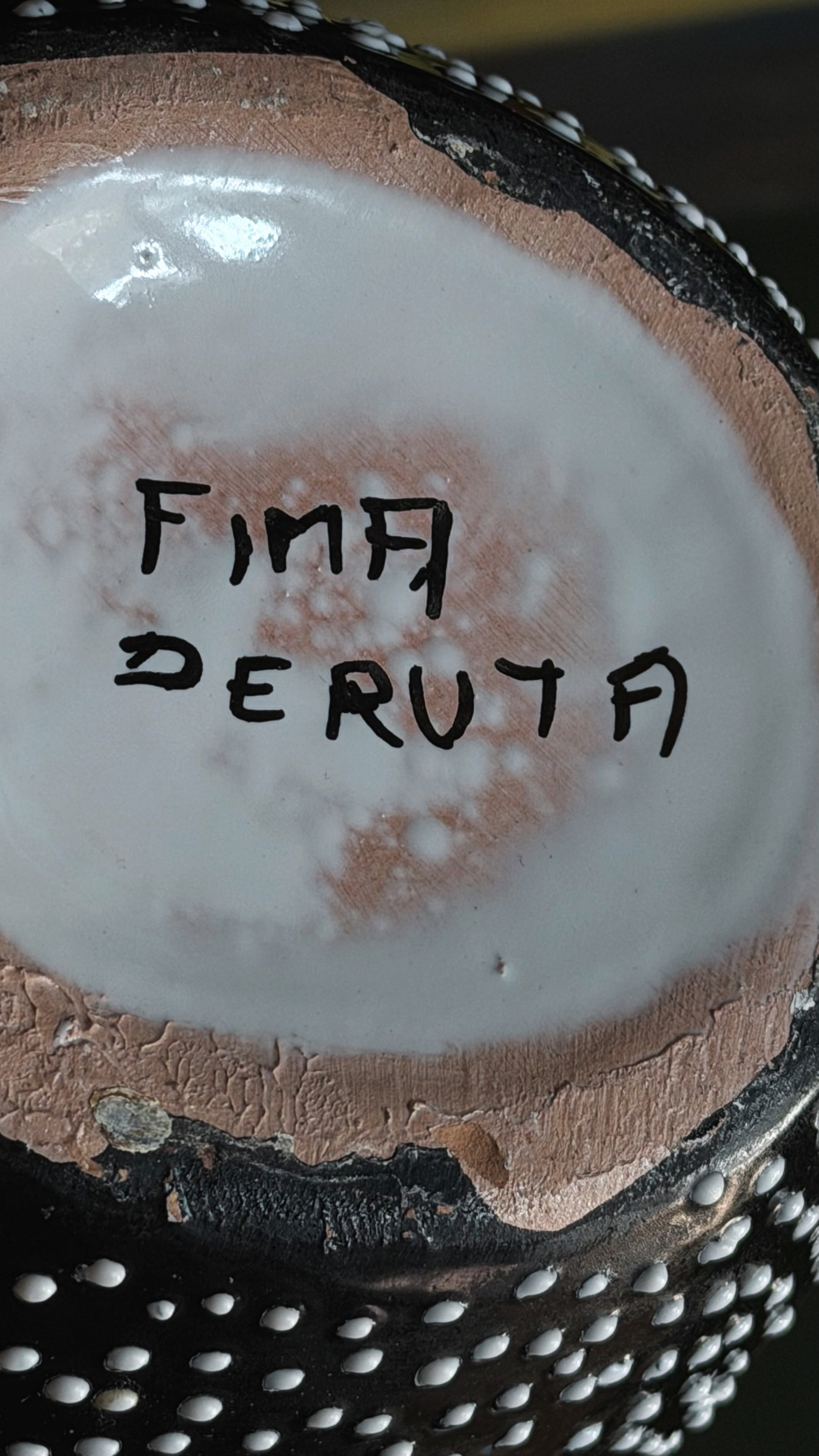 Close-up of the bottom of a handmade ceramic bowl or cup, showing a white glazed interior with the brand name 'FIMA DERUTA' written in black marker, surrounded by a rustic terracotta-colored rim and a black exterior with silver metallic studs.