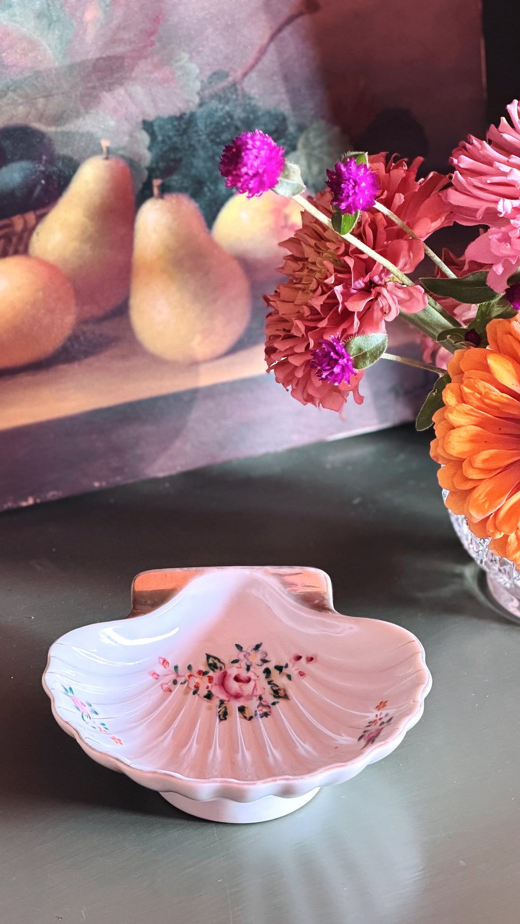 A pink ceramic clamshell-shaped dish with a floral design, set on a dark tabletop next to a glass vase of colorful flowers, with a still life painting of pears in the background.
