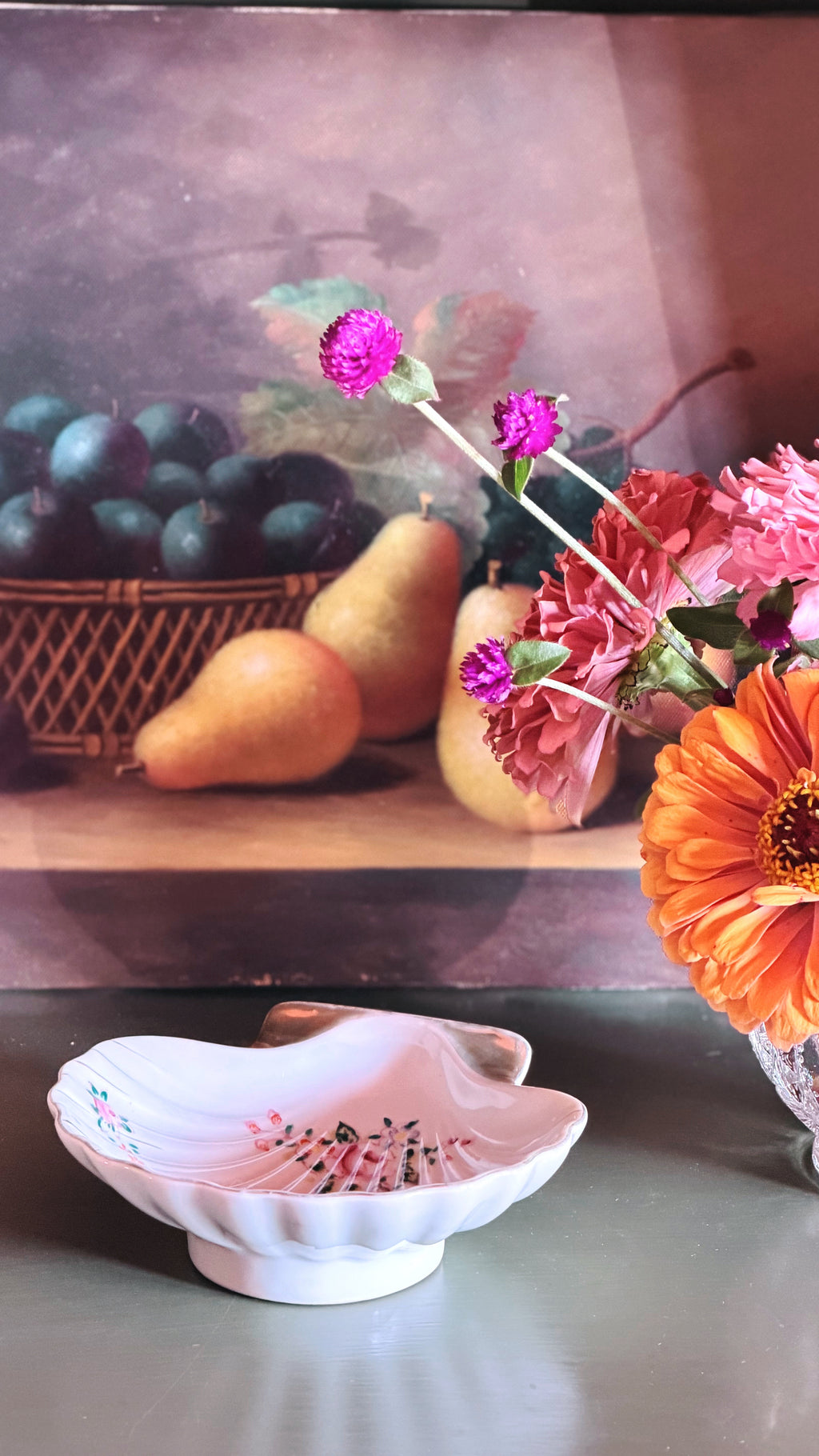 A white ceramic dish with a scalloped edge and delicate pink floral detailing, placed in the foreground on a tabletop. Behind it is a still life painting featuring pears, dark grapes, and flowers, including a prominent orange daisy and pink blooms.