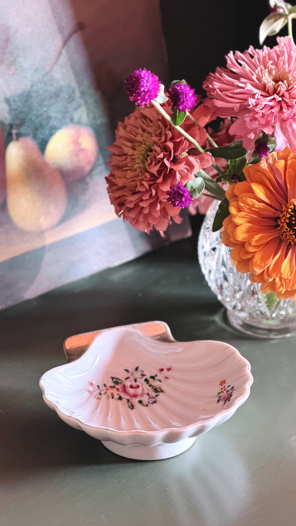 A small scalloped ceramic dish with a pink floral pattern, styled on a dark tabletop next to a glass vase of colorful flowers.