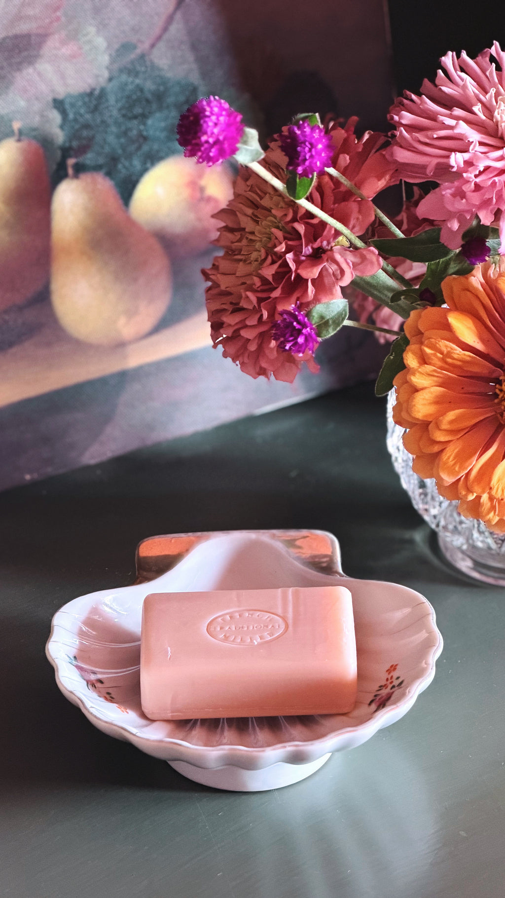 A pink rectangular bar of soap with an embossed circular logo, resting in a white scalloped dish with floral detailing. The dish is placed on a dark tabletop next to a glass vase filled with pink, orange, and purple flowers, with a blurred fruit painting in the background.