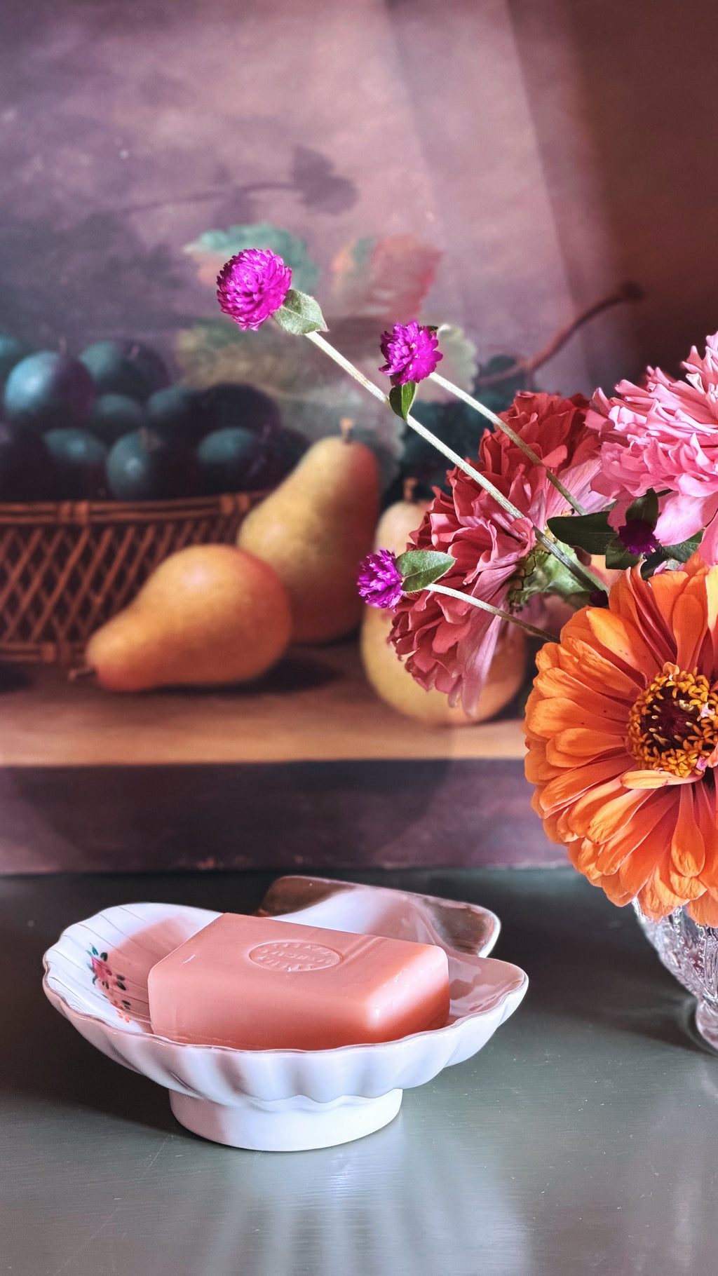 A pink rectangular bar of soap sits in a white scalloped dish on a tabletop, with a floral arrangement and fruit in the softly blurred background.