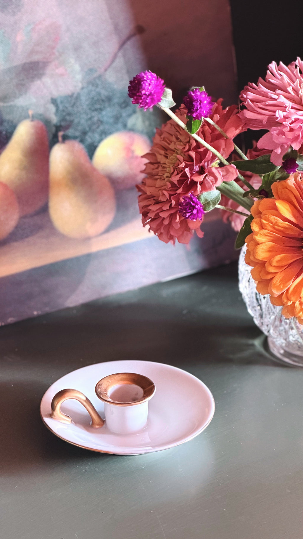 A small white ceramic candleholder with a metallic rose gold rim and handle, resting on a matching saucer, set on a dark tabletop next to a glass vase filled with pink, orange, and purple flowers.