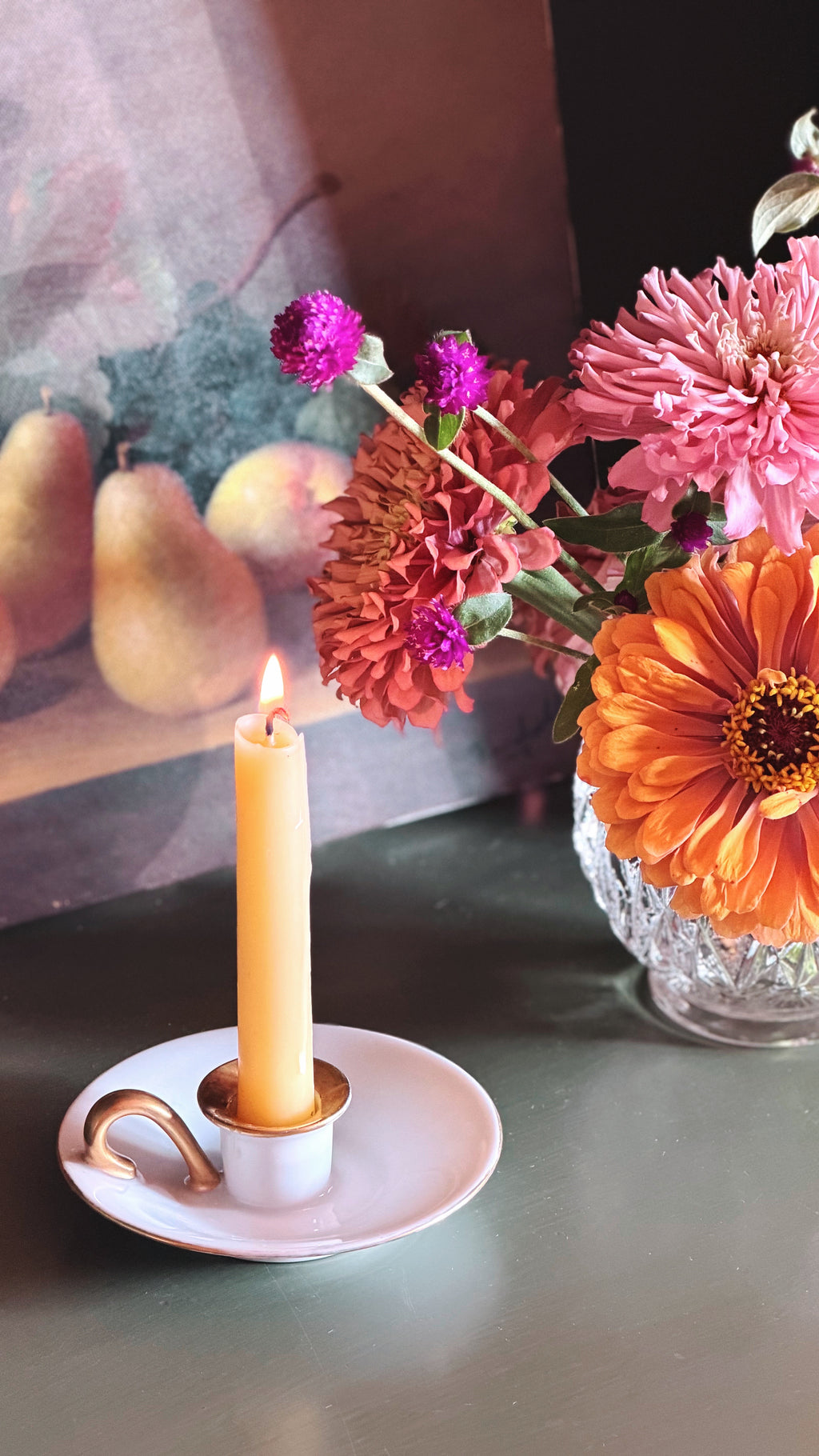 A lit yellow taper candle in a small white holder with a gold accent, set on a white saucer with a gold handle, next to a glass vase filled with pink, orange, and purple flowers.