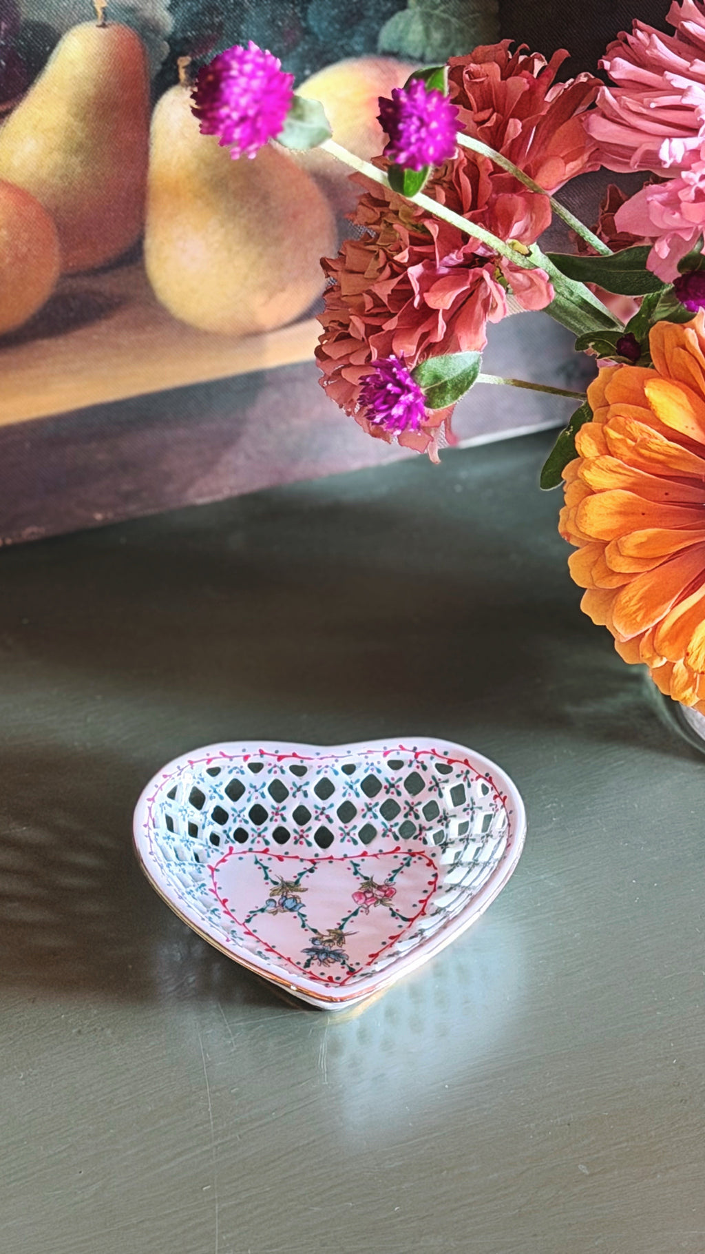 A small heart-shaped ceramic dish with a white lattice pattern and pink floral accents, placed on a dark tabletop with flowers and pears in the background.