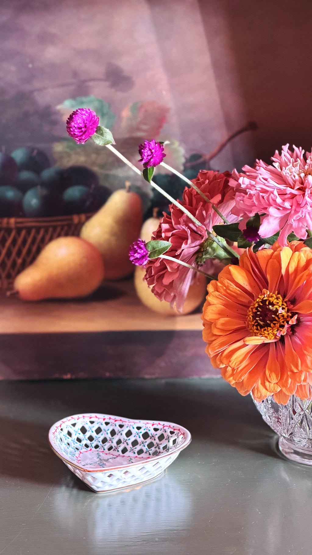 A still life arrangement with a vase of colorful flowers, including a prominent orange zinnia and pink and purple blooms, alongside a small white heart-shaped lace dish on a tabletop.