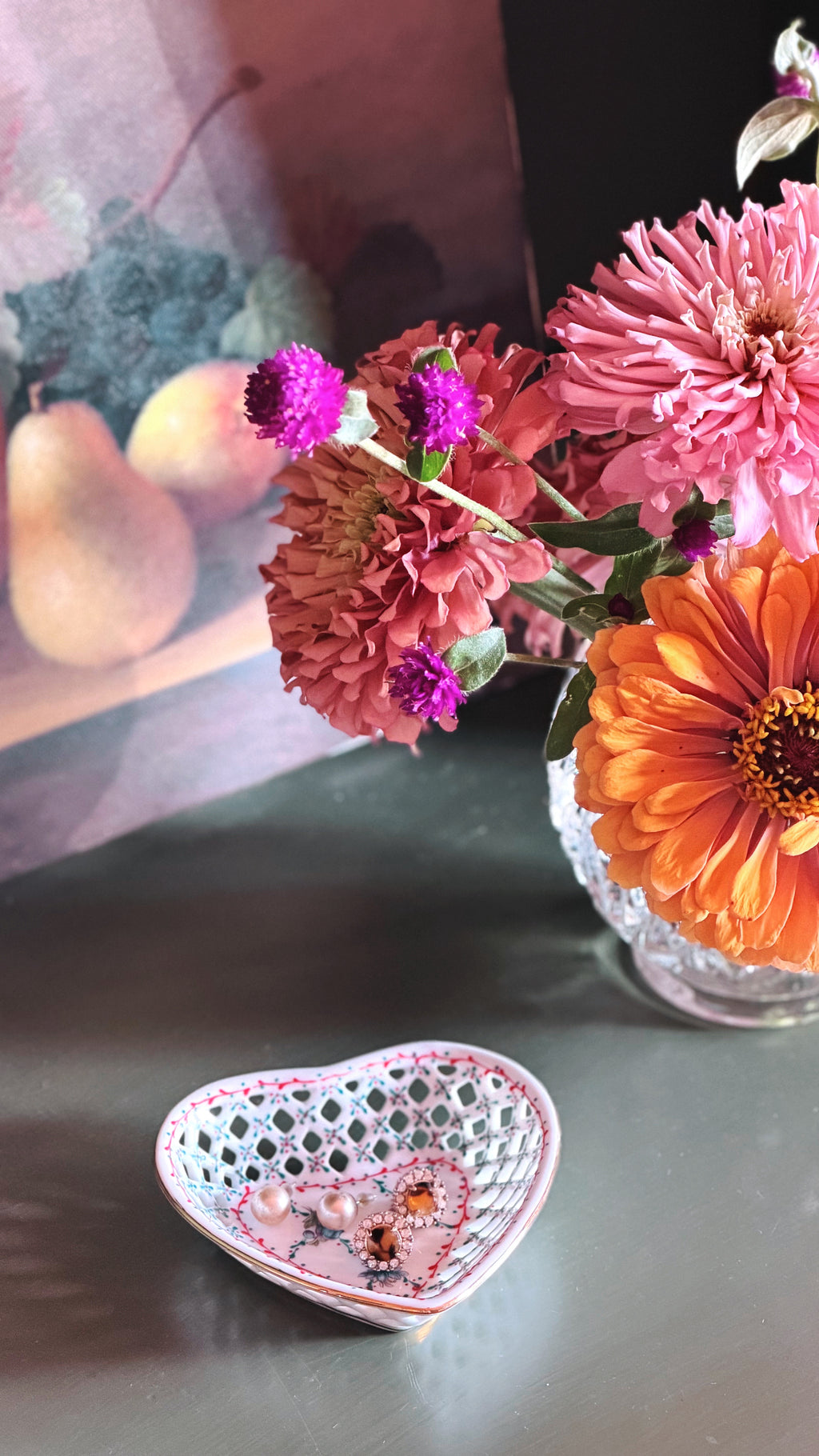 A heart-shaped white ceramic dish with pink floral patterns and a white lattice cutout, holding small jewelry pieces, set on a dark tabletop next to a glass vase of colorful flowers.