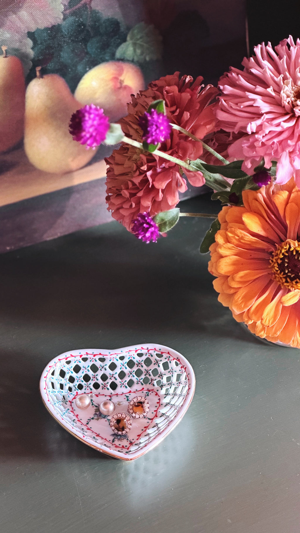 A heart-shaped ceramic jewelry box with a white lattice top and pink floral detailing, containing earrings, placed on a dark tabletop next to a colorful flower arrangement.