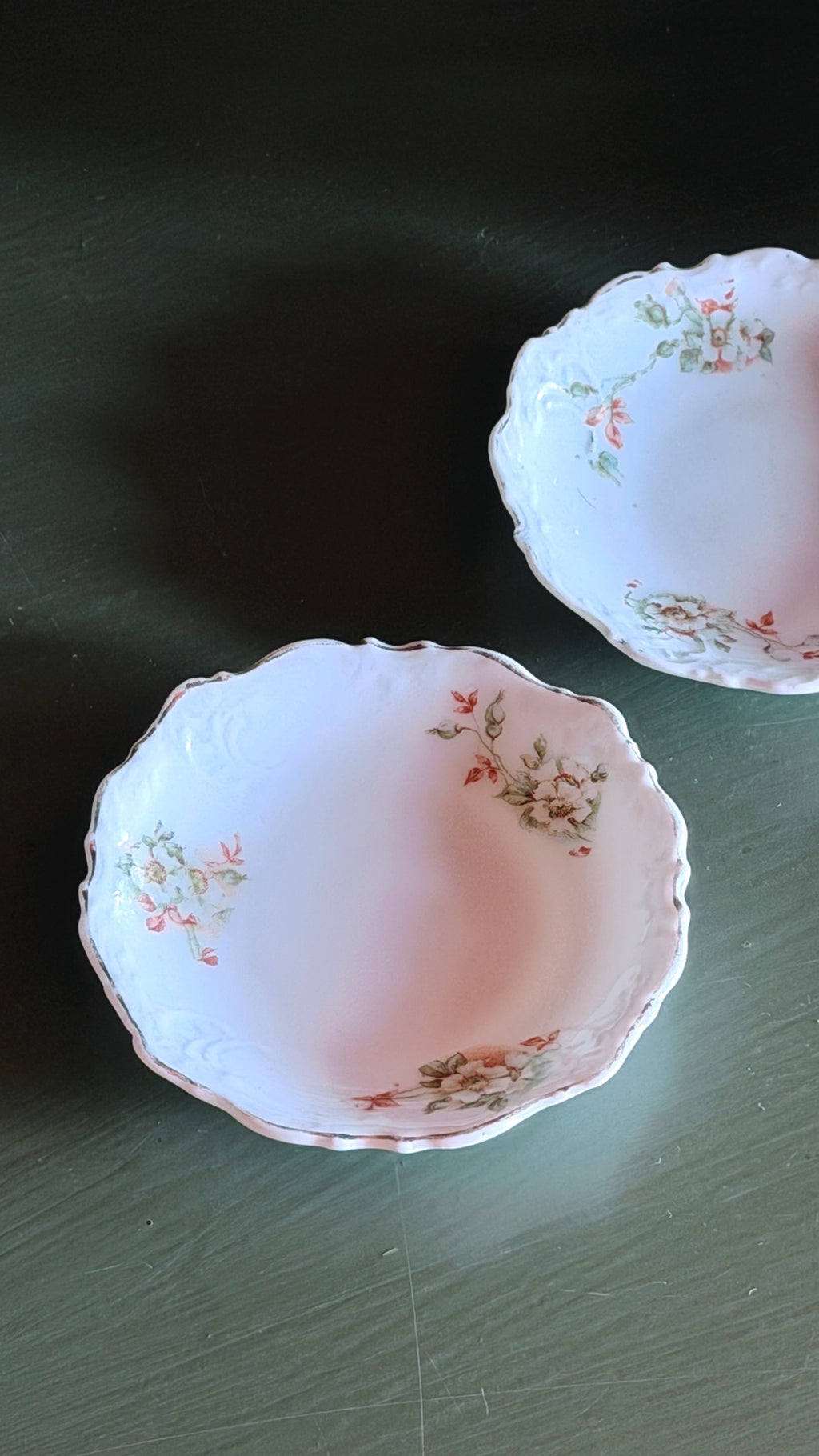 Two white ceramic bowls with scalloped edges and delicate pink and green floral patterns, displayed on a dark green surface.