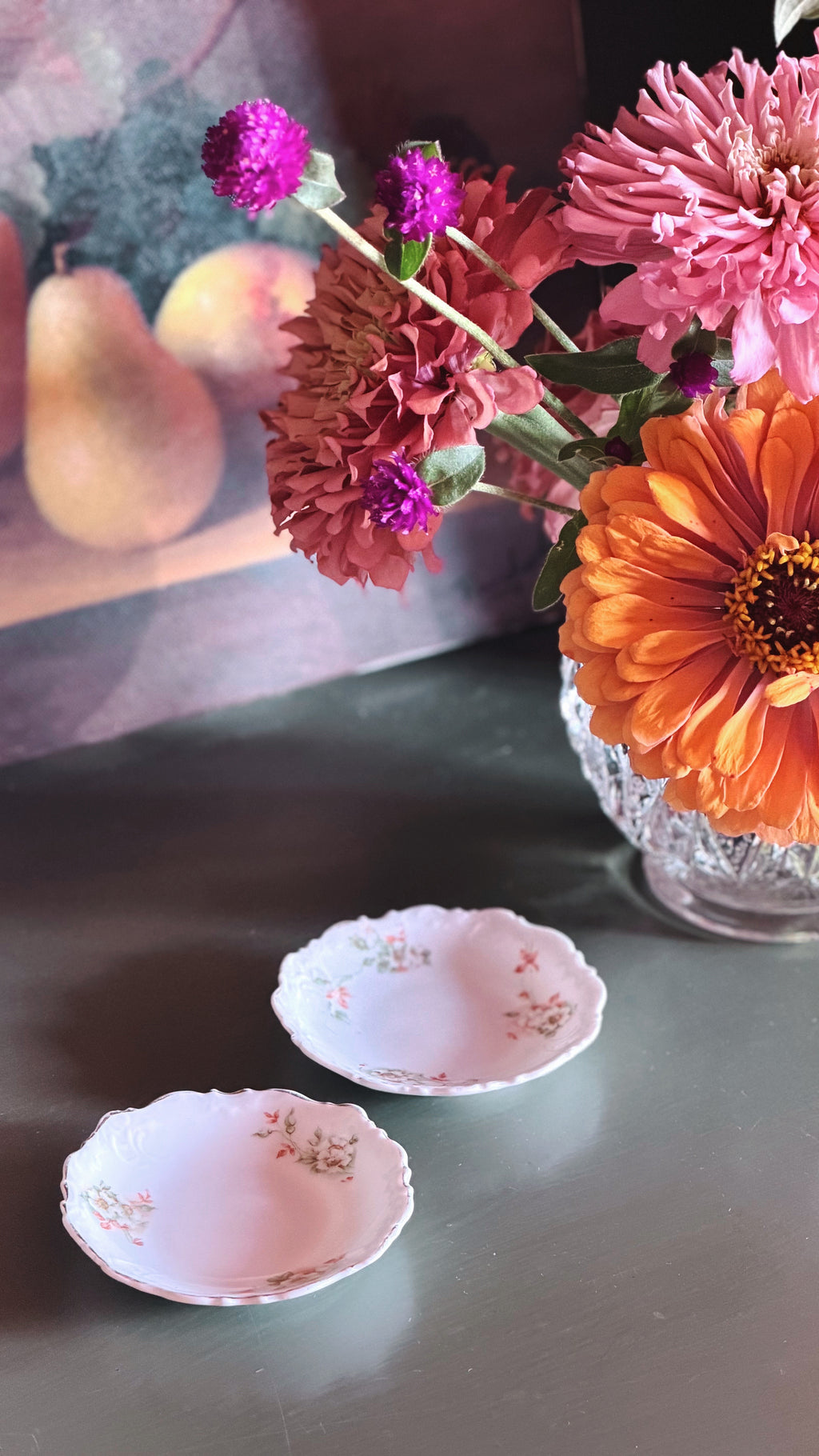 Two small white ceramic plates with delicate pink floral patterns and scalloped edges, arranged on a dark tabletop next to a glass vase filled with colorful flowers.