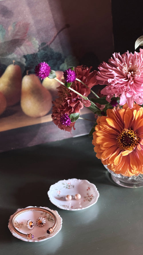 A styled tabletop scene with a bouquet of pink, orange, and purple flowers in a glass vase, alongside two small floral jewelry stands holding earrings and rings. The background includes a painted pear still life and a dark surface.