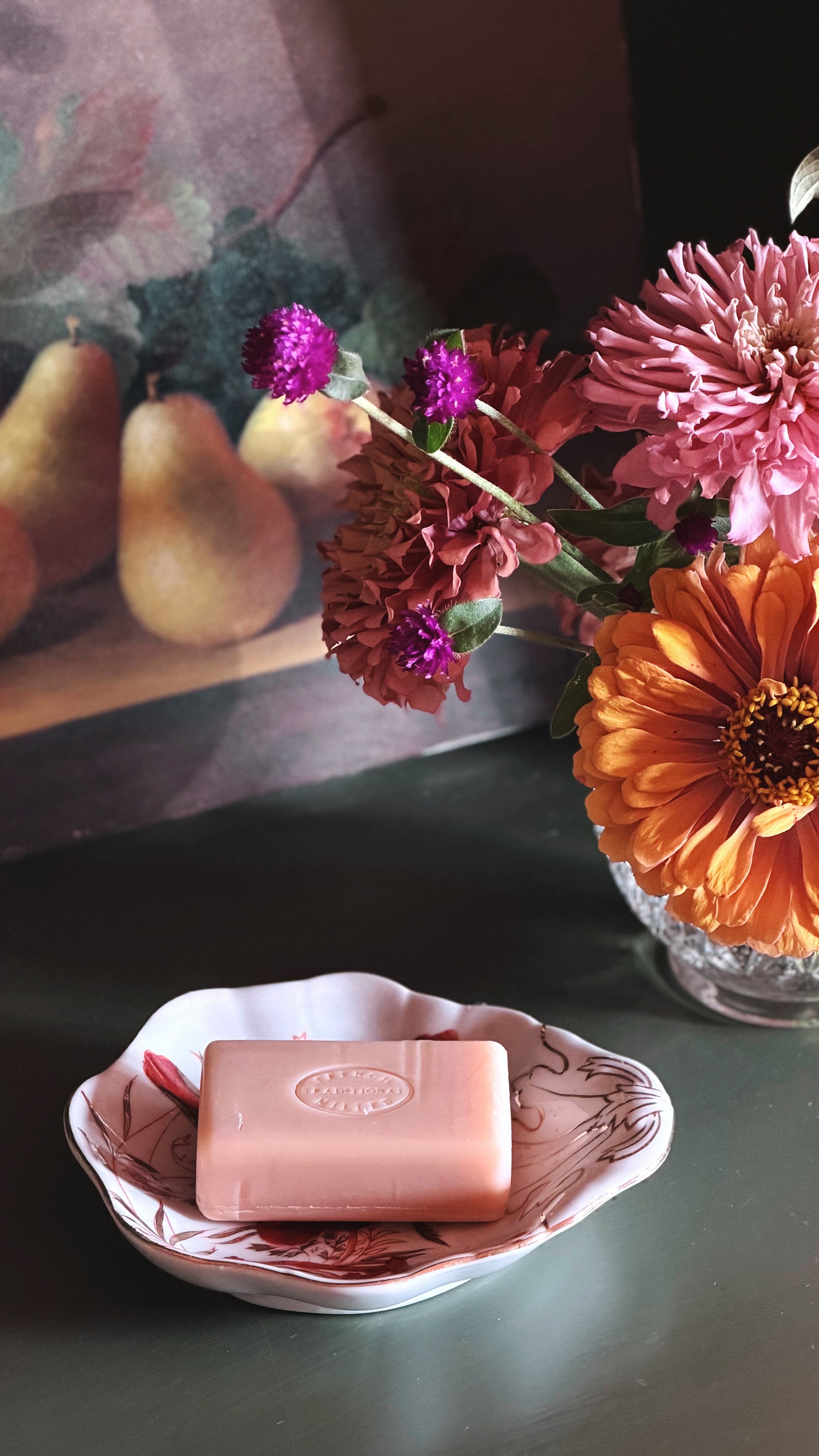 A pink rectangular bar of soap rests in a small white dish with a floral pattern, set on a dark tabletop. In the background, a glass vase holds orange and pink flowers, and a framed still life painting of pears is visible.