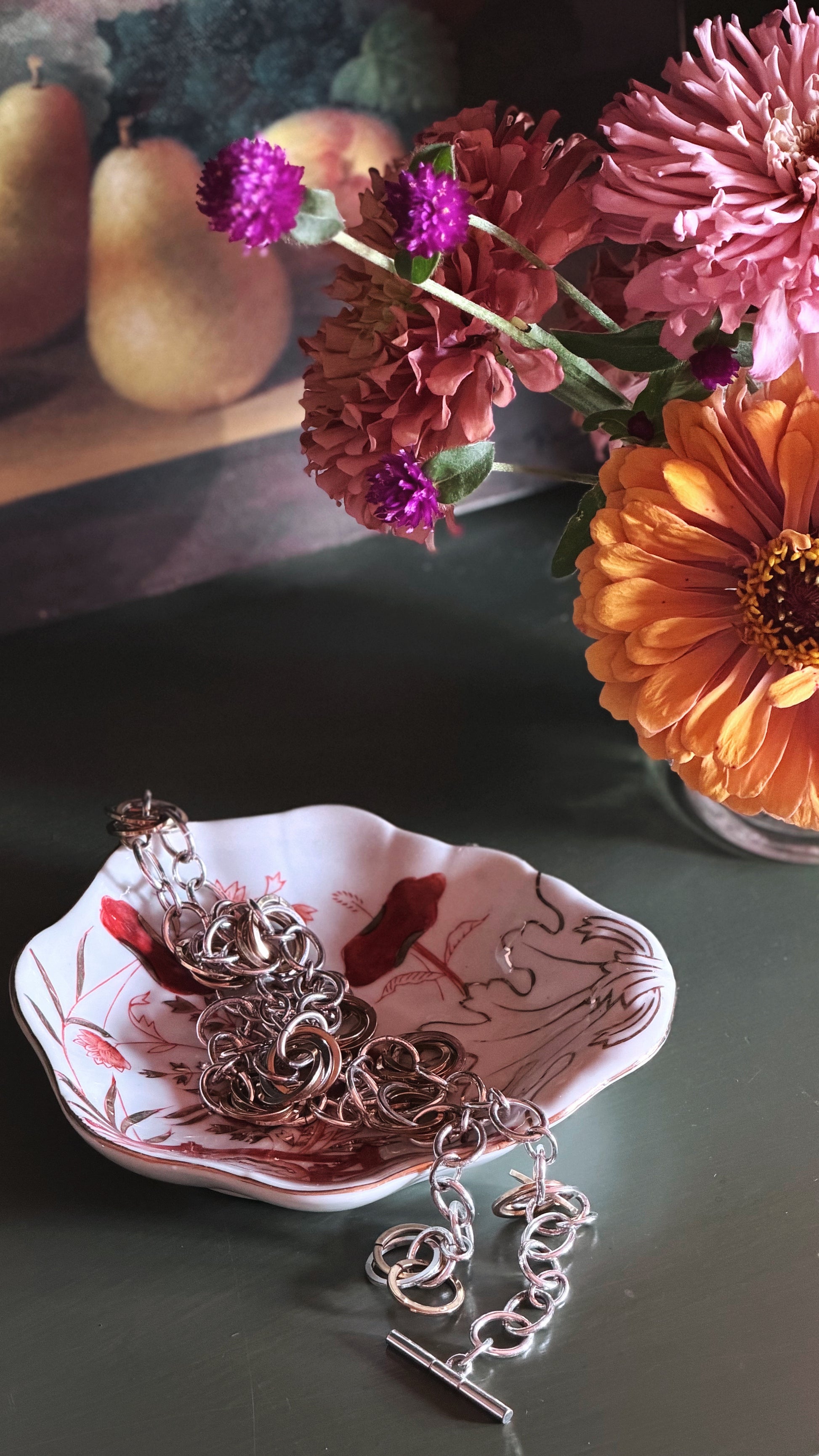 A silver-toned chain necklace with a toggle clasp is draped over a small decorative dish with a pink and white floral pattern, set on a dark tabletop next to a vase of colorful flowers.
