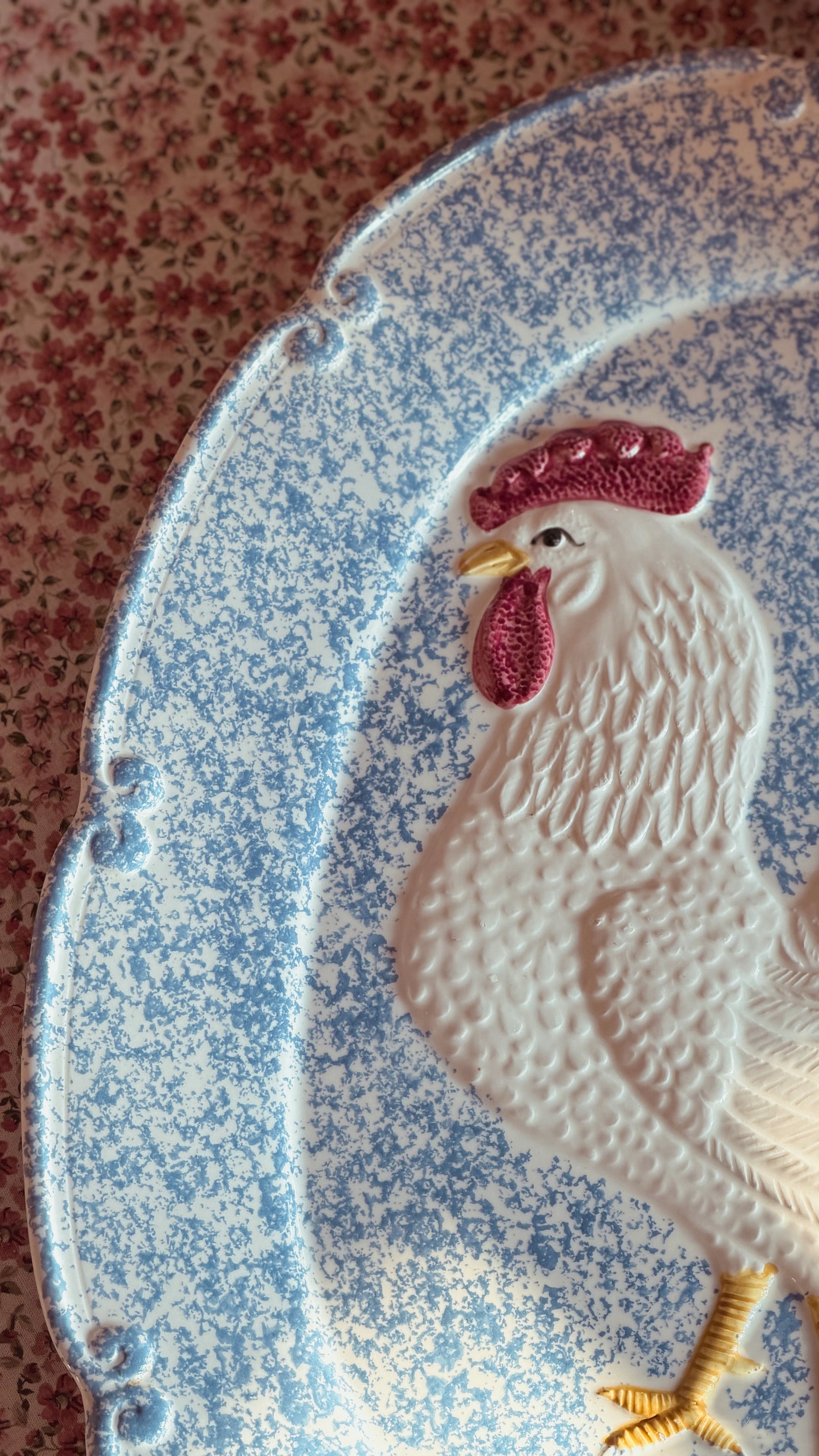 A close-up of a decorative ceramic platter featuring a raised white rooster with a pink comb and yellow beak and leg, set against a blue speckled background.
