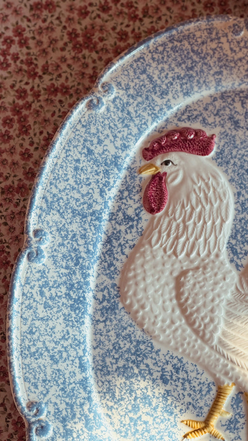 A close-up of a decorative ceramic platter featuring a raised white rooster with a pink comb and yellow beak and leg, set against a blue speckled background.