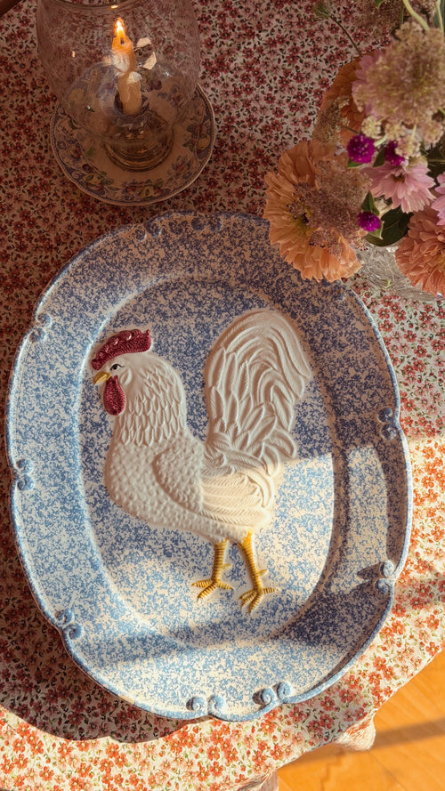 An oval blue and white speckled ceramic platter featuring a raised white rooster design with a red comb and yellow legs, set on a floral tablecloth with a candle and flowers nearby.