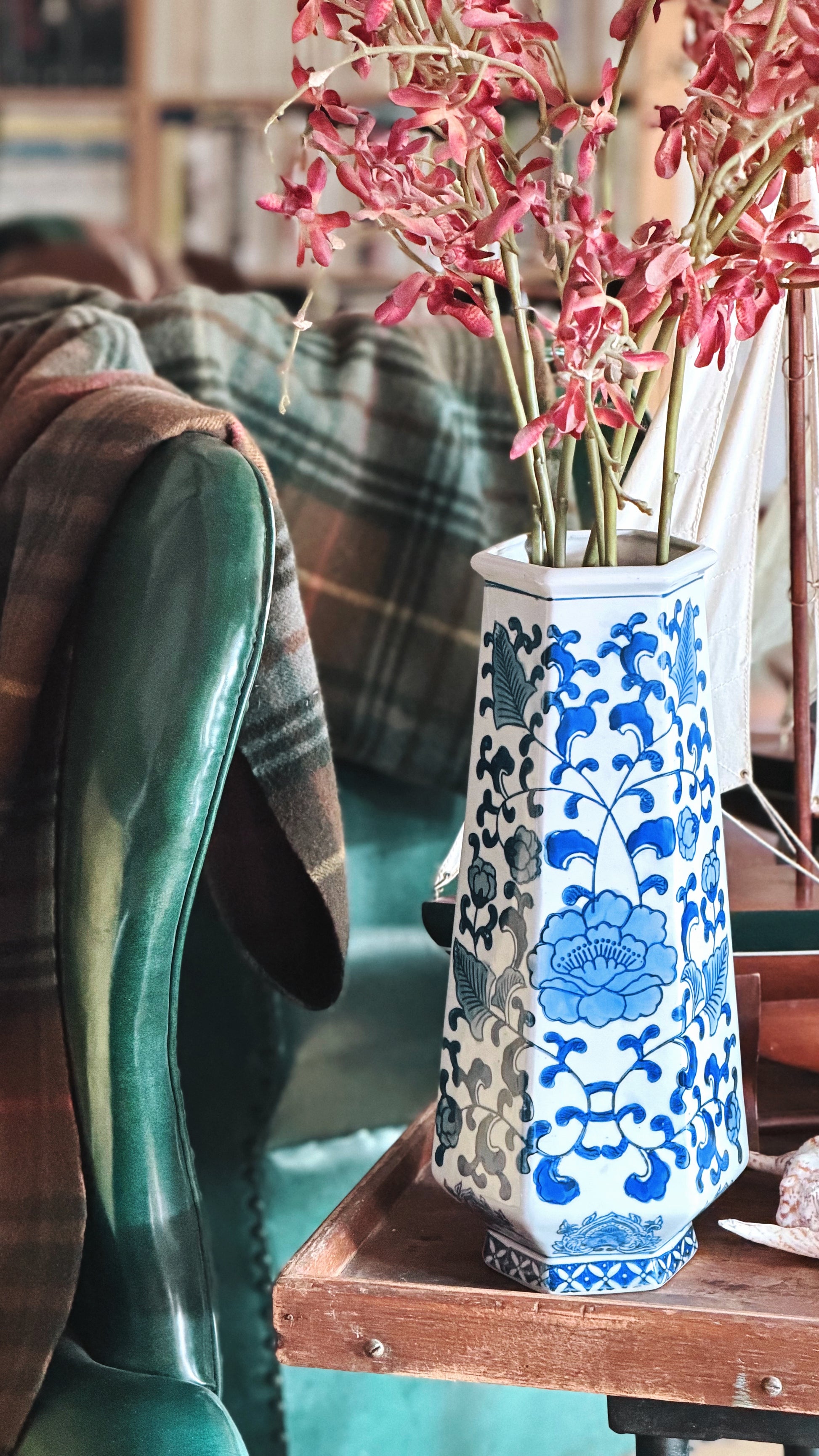 A tall white ceramic vase with blue floral patterns, filled with pink dried flowers, sitting on a wooden table in a cozy indoor setting next to a green leather chair.