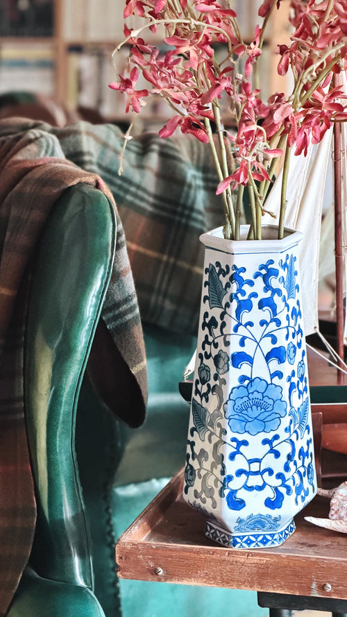 A tall white ceramic vase with blue floral patterns, filled with pink dried flowers, sitting on a wooden table in a cozy indoor setting next to a green leather chair.