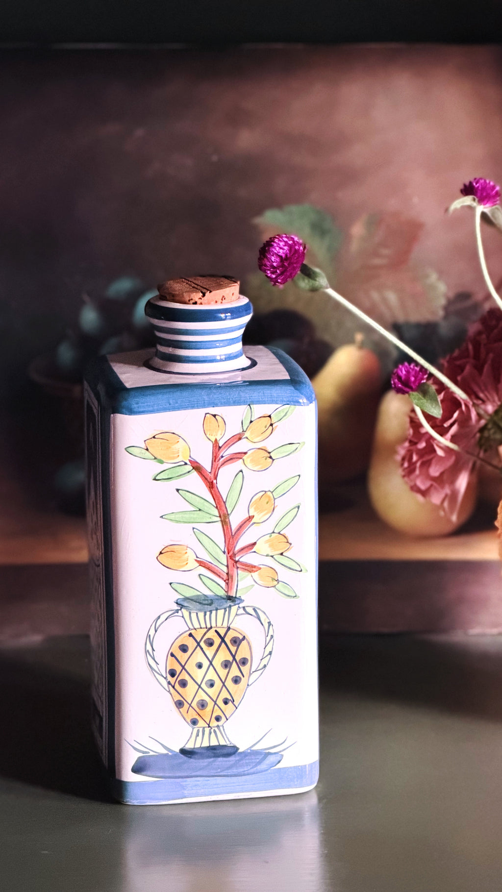 A rectangular ceramic bottle with a cork stopper, decorated with a hand-painted floral design of yellow flowers in a patterned vase. The bottle features a white body with blue stripes on the neck and base, and is set against a dark, rustic background with blurred dried flowers.