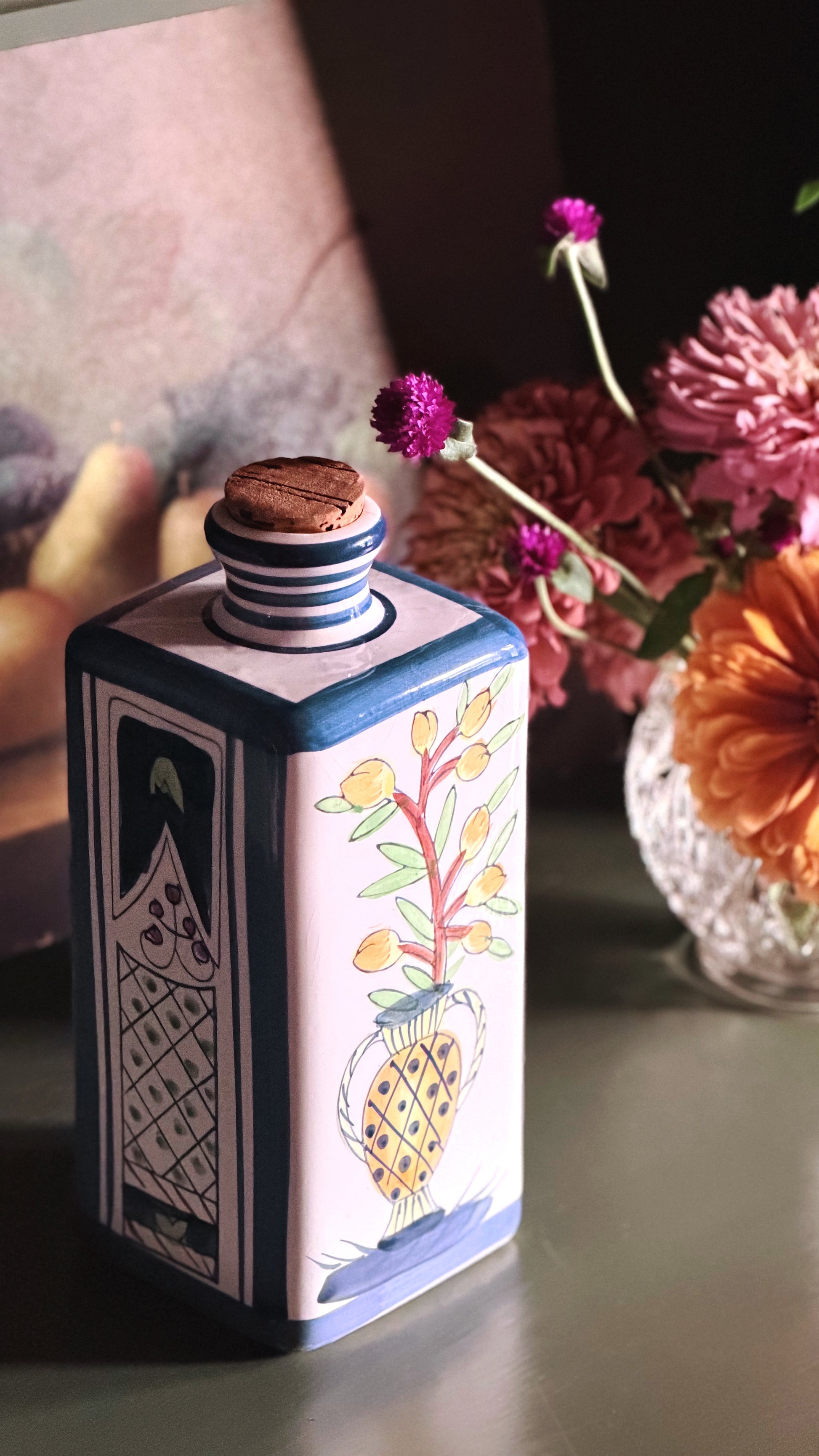 A square decorative ceramic bottle with a cork stopper, featuring hand-painted floral and geometric designs in blue, white, yellow, and green, set against a warm, softly blurred background with flowers.