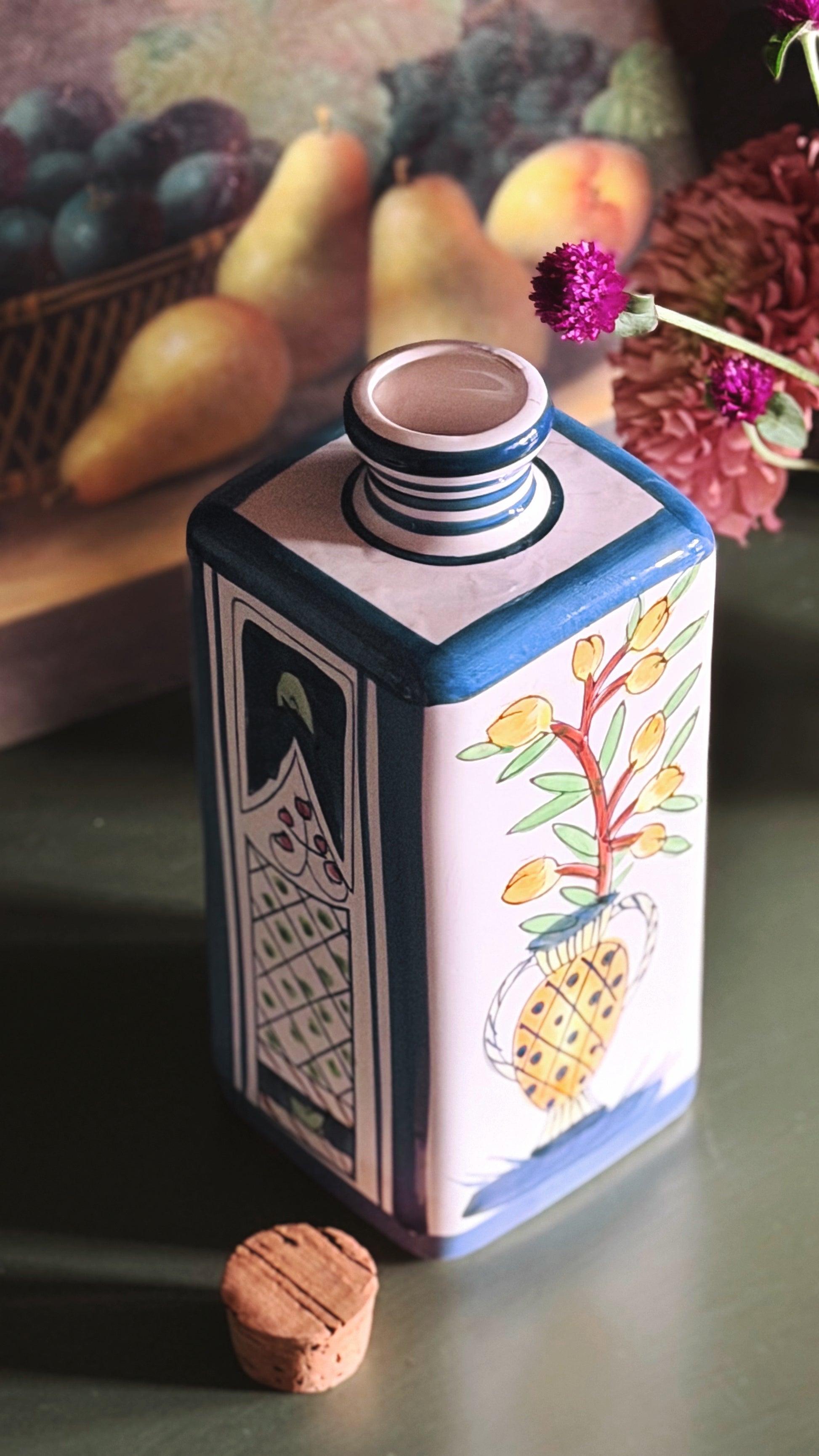 A hand-painted ceramic bottle with a cork stopper, featuring a white body decorated with blue borders, a fish illustration, and a yellow grapevine motif, set against a dark background with blurred fruit and flowers.