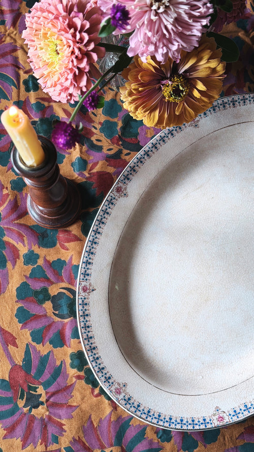An oval serving platter with a blue and white patterned rim, placed on a colorful floral tablecloth next to a lit candle and flowers.