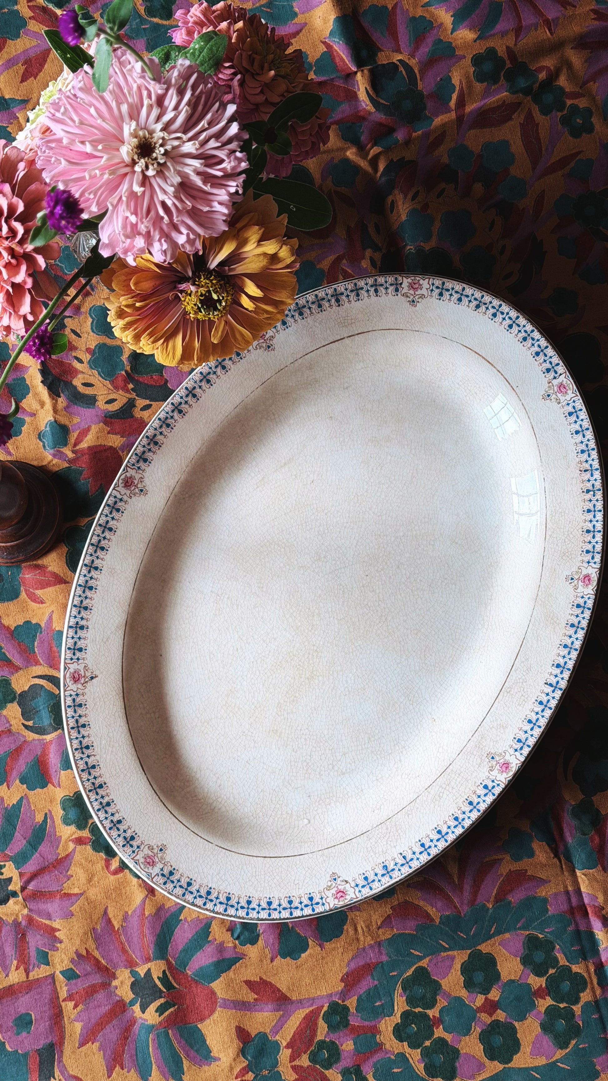 An oval serving platter with a blue and pink floral border, displayed on a vibrant, multicolored floral-patterned tablecloth next to pink and yellow flowers.