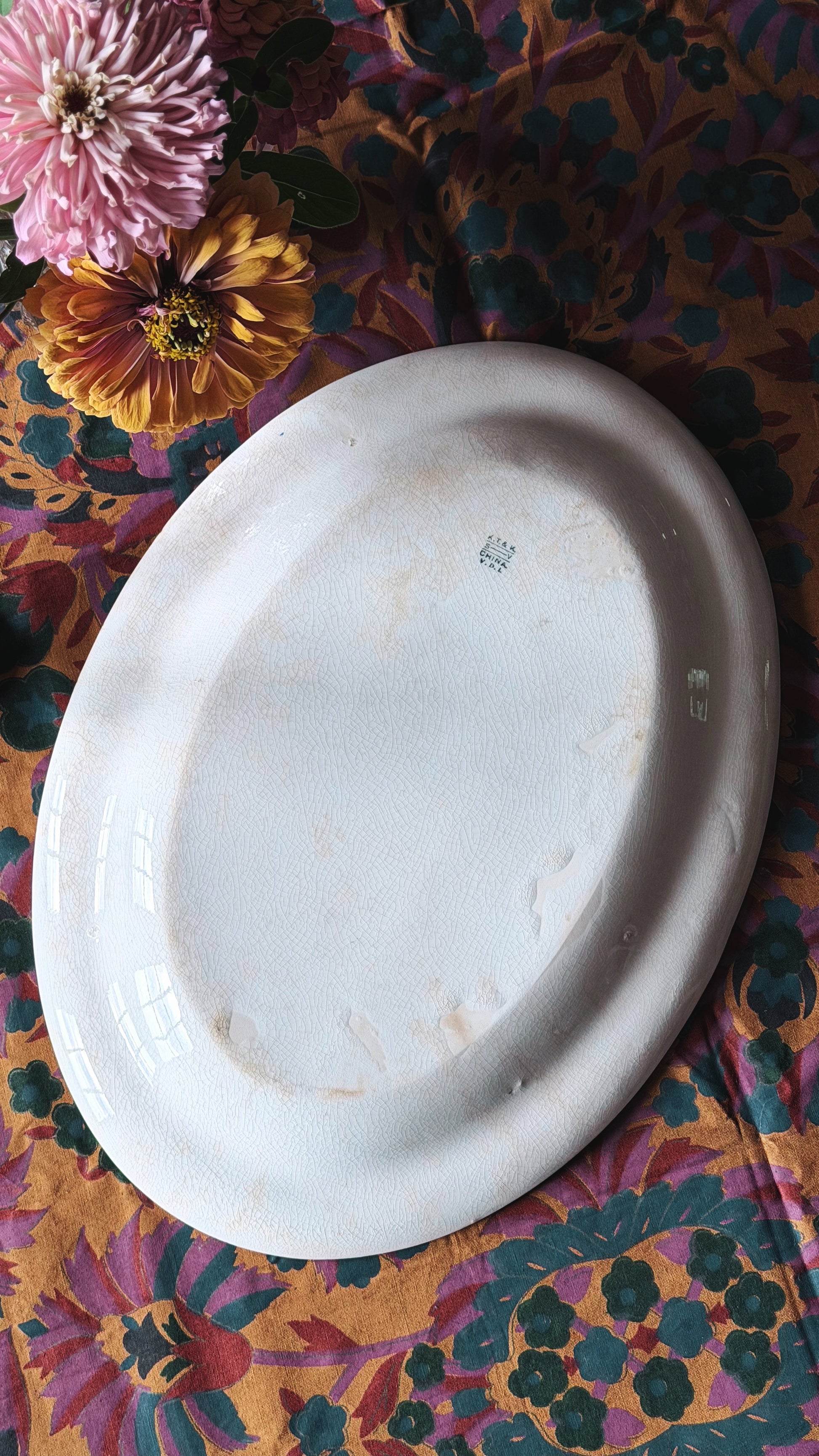 An oval white platter with a glossy glaze, shown from a top-down view on a colorful floral patterned fabric with pink and yellow flowers.