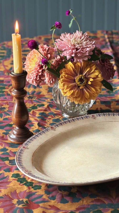A styled tabletop scene with a Vintage Knowles Taylor Knowles Oval Serving Platter in the foreground, a lit candle in a dark wooden holder, and a glass vase holding pink and yellow flowers, all set on a colorful floral tablecloth.