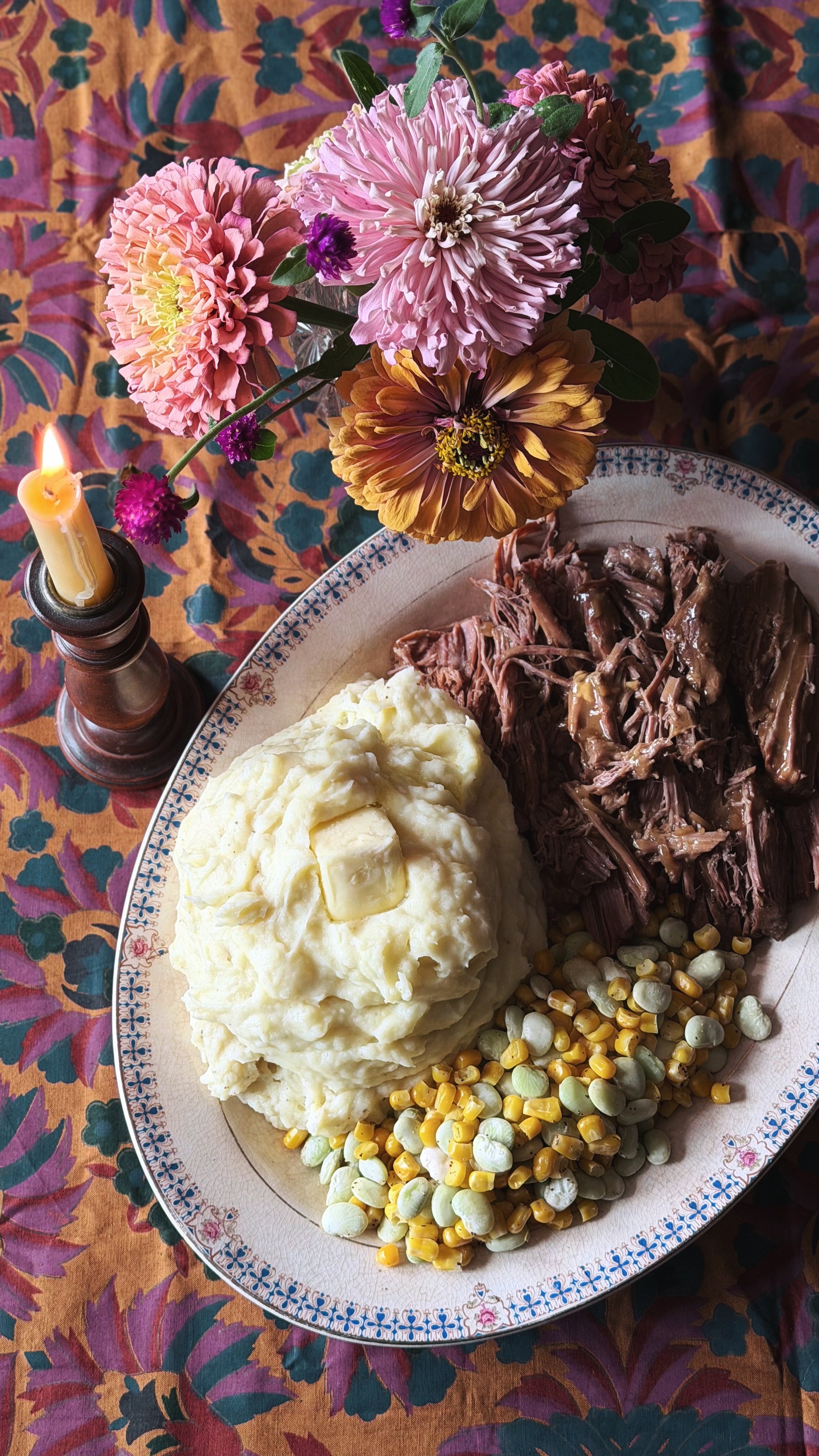 A rustic plate of mashed potatoes, shredded beef, and mixed beans on a patterned tablecloth, styled with a candle and flowers.