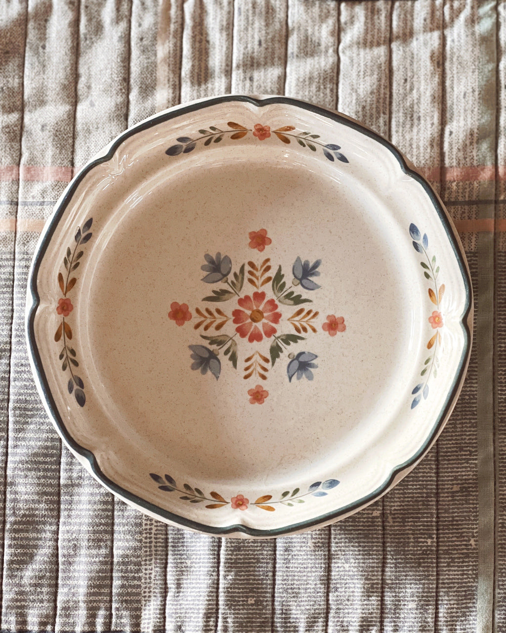 A International Tableworks American Patchwork Salad Plate with a scalloped rim, featuring a central floral medallion and delicate leafy accents in muted orange, blue-gray, and olive green, set against a textured beige and brown striped fabric background.