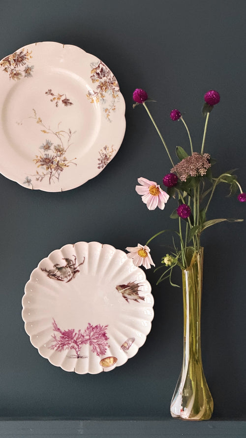 Two decorative ceramic plates with floral and bird illustrations are displayed against a dark gray wall, alongside a tall gold glass vase holding pink and purple flowers.