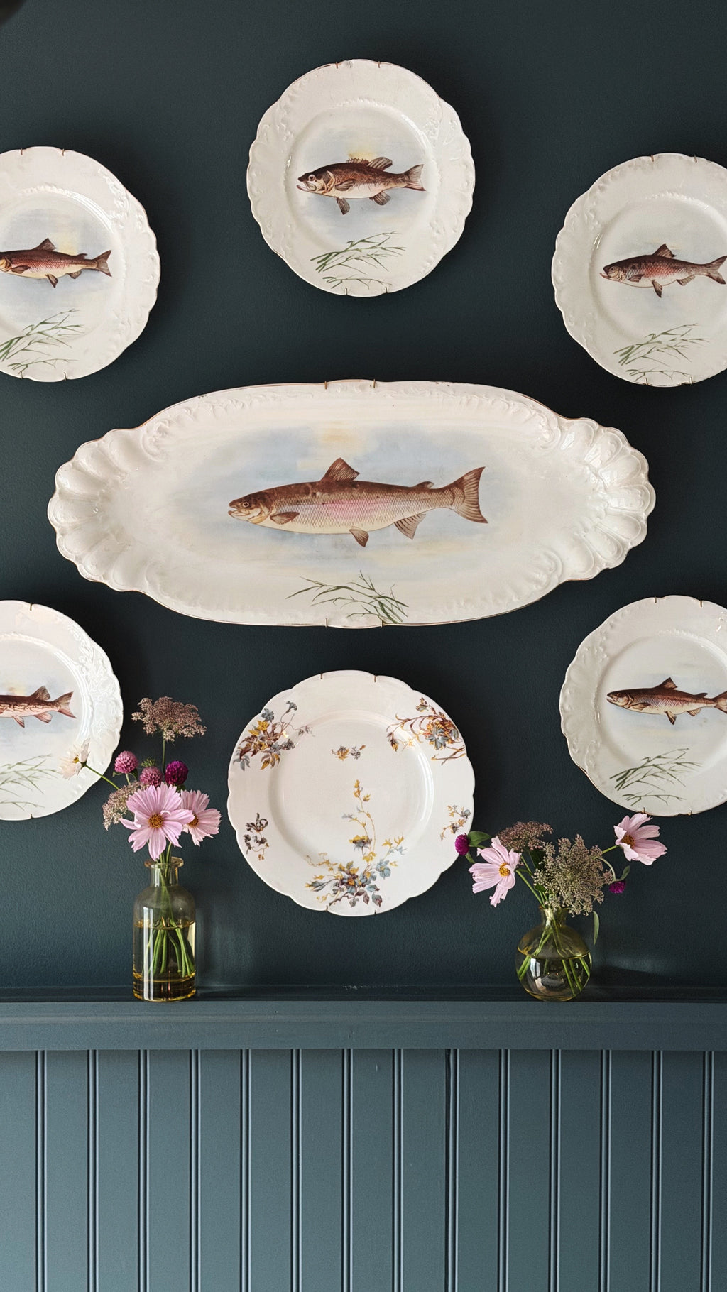 A set of white ceramic plates with scalloped edges and hand-painted fish illustrations, displayed on a dark teal wall above a matching paneled shelf with small pink flower arrangements.
