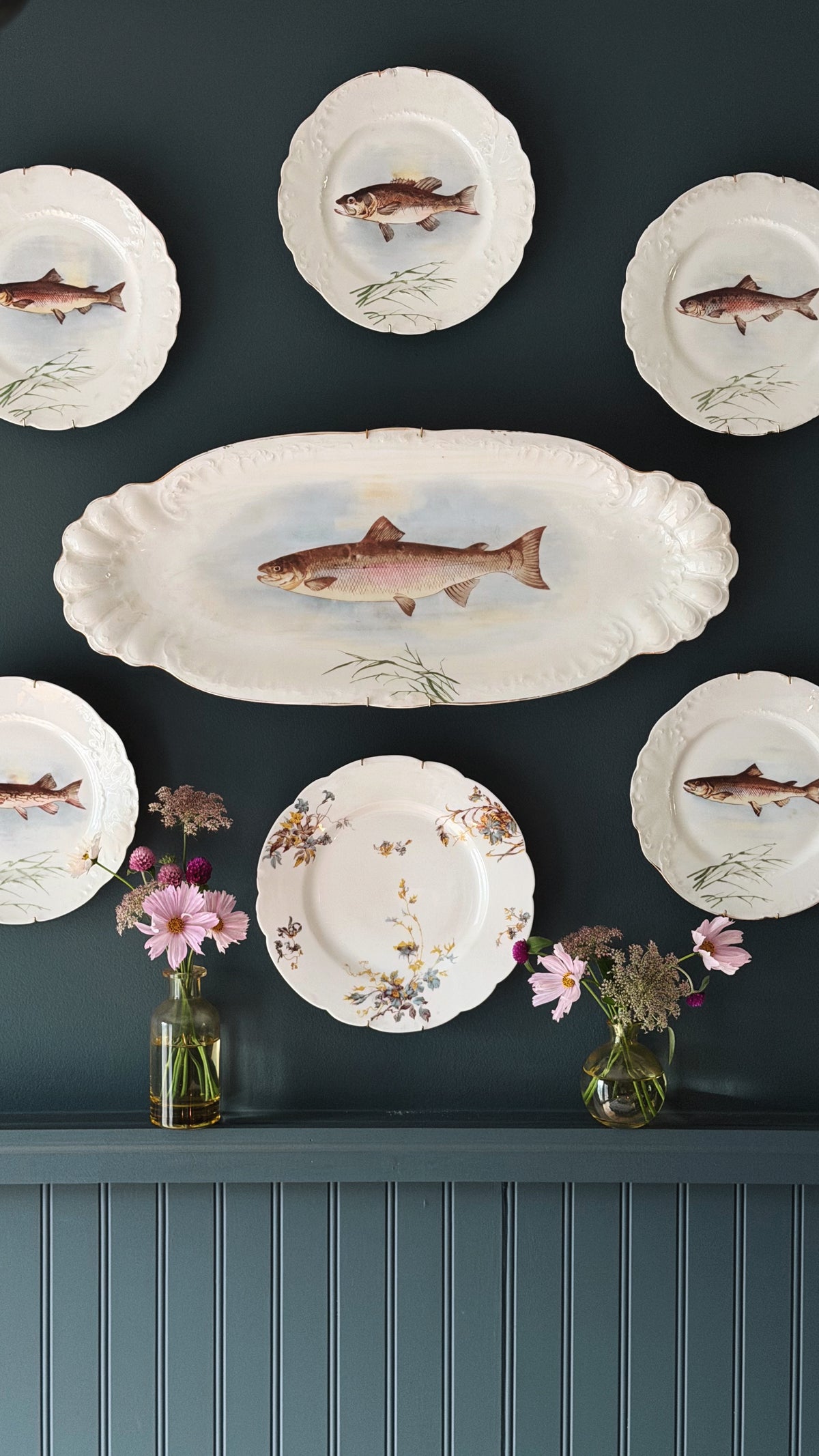 A set of white ceramic plates with scalloped edges and hand-painted fish illustrations, displayed on a dark teal wall above a matching paneled shelf with small pink flower arrangements.