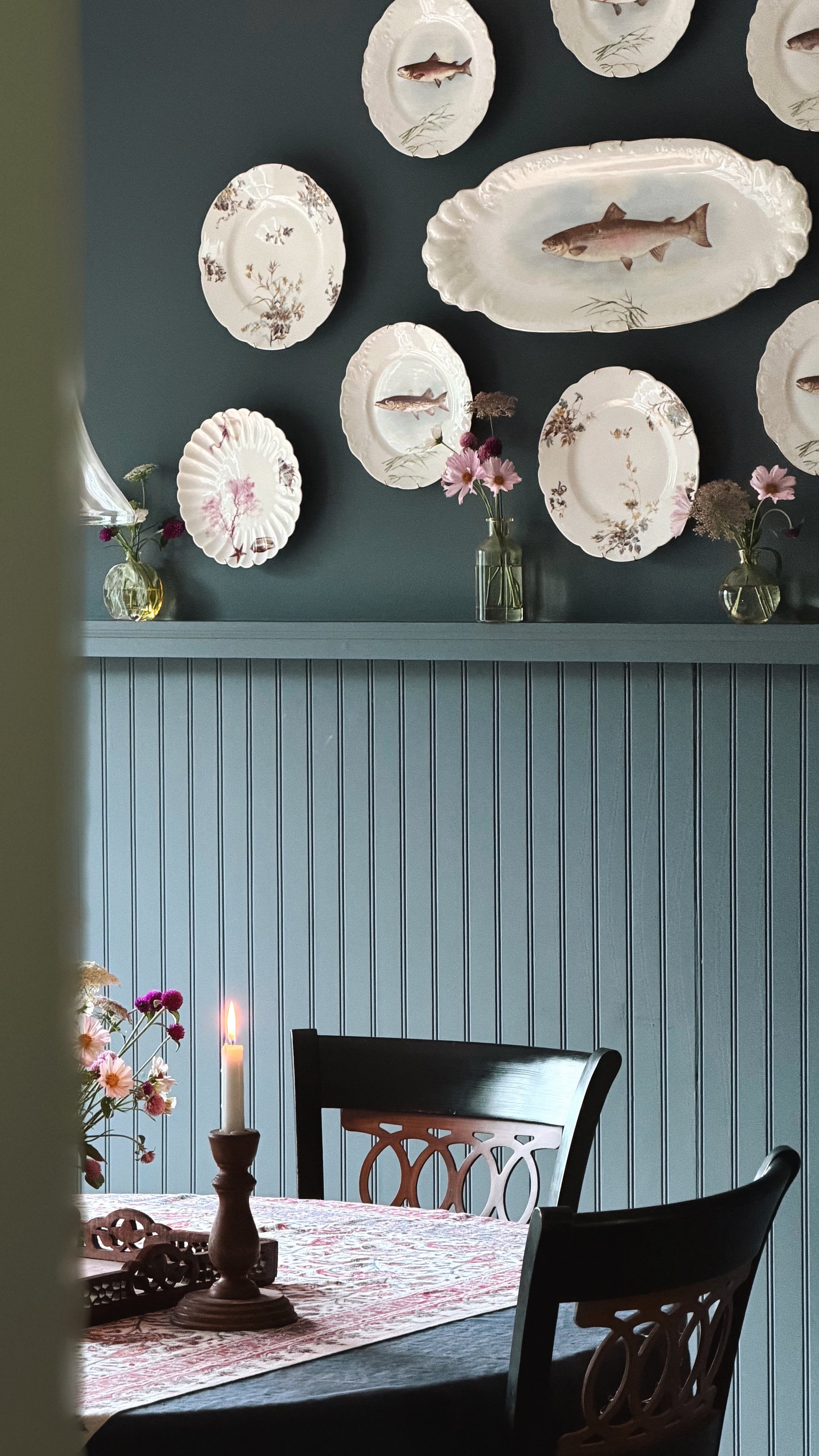 A styled dining room corner with a dark teal wall decorated with vintage white plates and fish-shaped serving platters, above a light blue paneled wall. A dining table with dark wooden chairs, a candlestick, and floral arrangements is visible in the foreground.