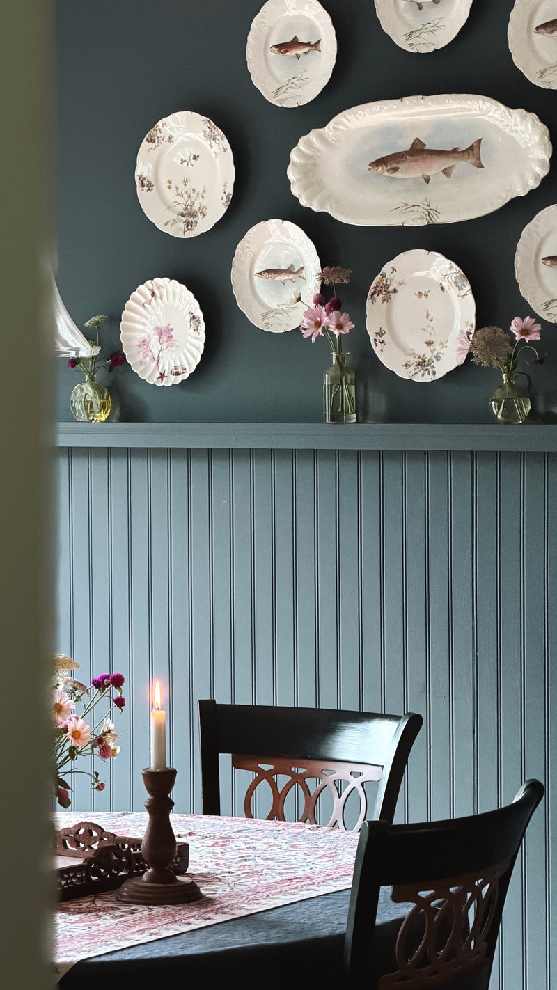 A styled dining room corner with a dark teal wall decorated with vintage white plates and fish-shaped serving platters, above a light blue paneled wall. A dining table with dark wooden chairs, a candlestick, and floral arrangements is visible in the foreground.