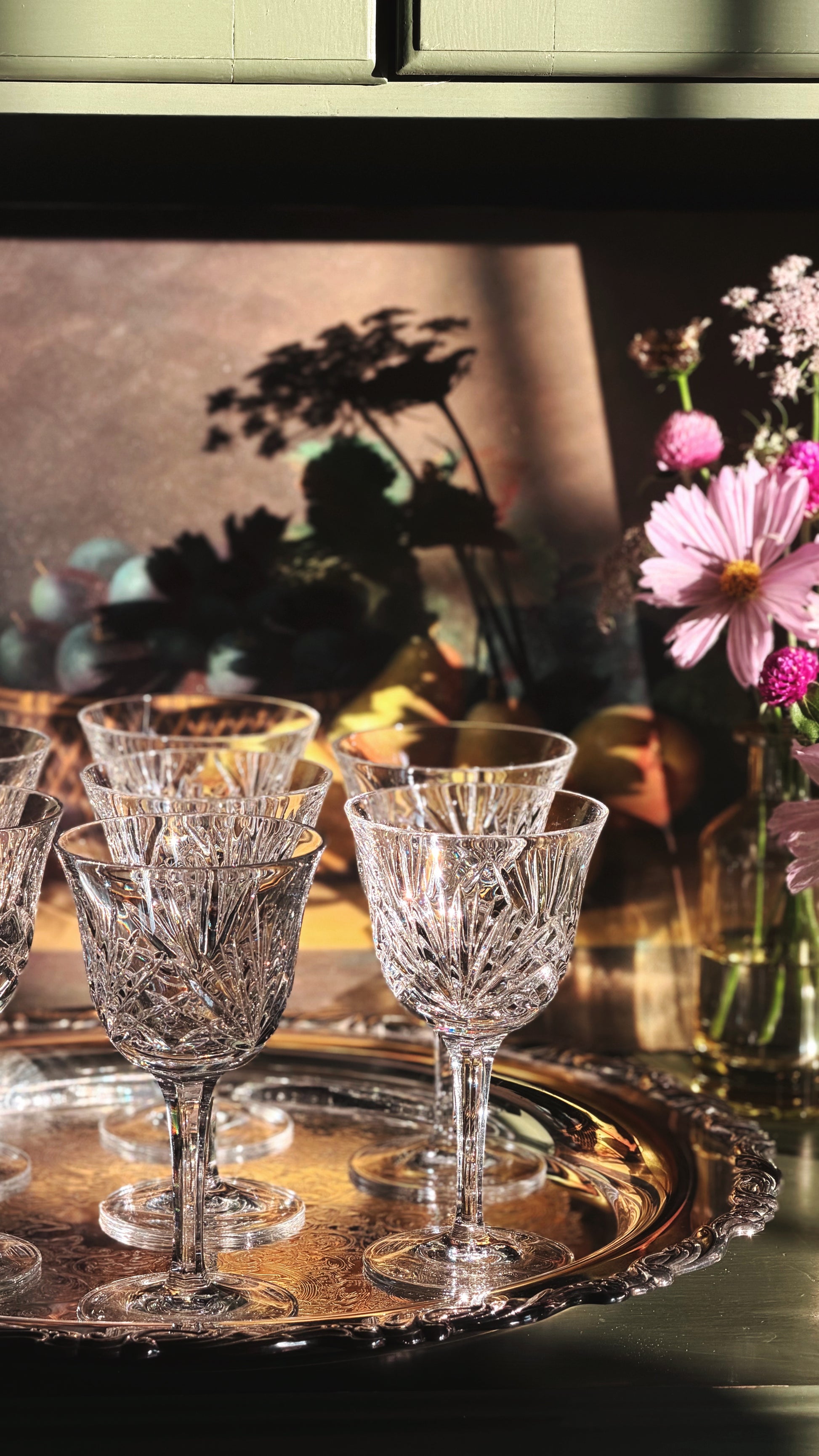A set of clear cut-crystal wine glasses arranged on a round silver tray, with a warm, softly lit background and pink flowers in a vase to the right.